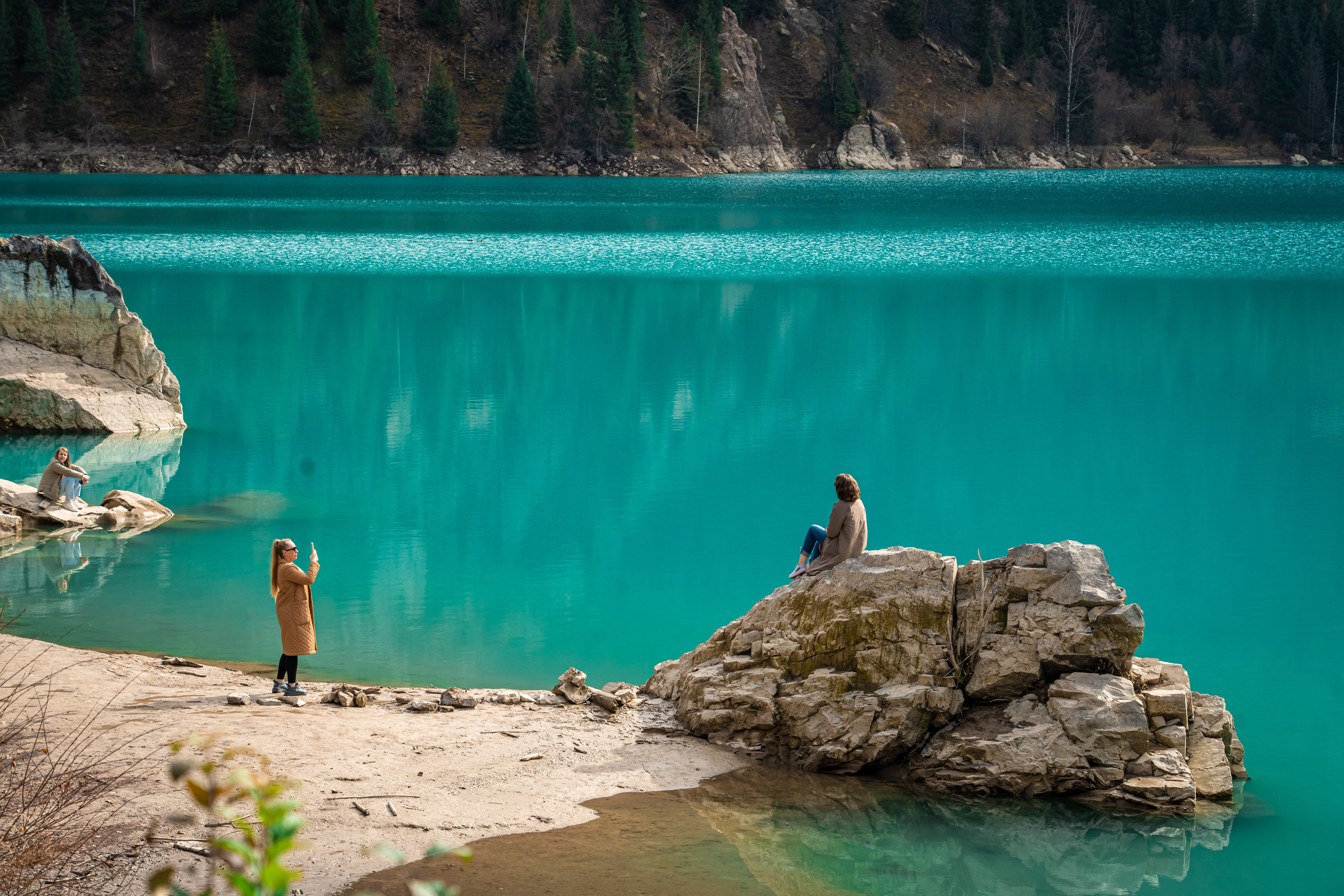 Вид на Лунный Каньон реки Чарын недалеко от Алматы, окружённый зелёными деревьями и кустарниками, на фоне красочных скал в солнечный день. На переднем плане женщина сидит на утёсе и любуется пейзажем