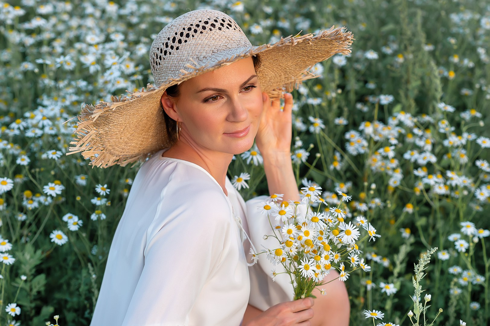 Красивая девушка в соломенной шляпе сидит в ромашковом поле и держит в руках букет ромашек