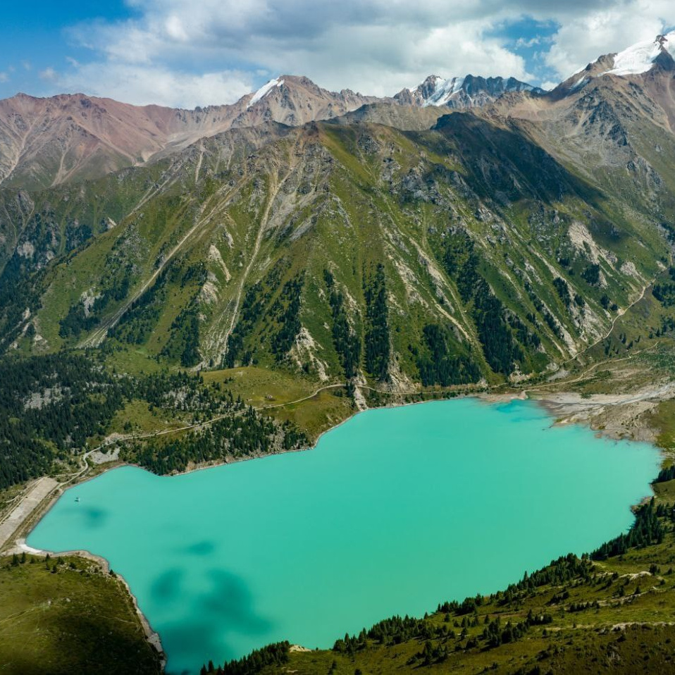 Ярко-бирюзовое озеро Иссык недалеко от Алматы окружено густыми хвойными лесами на фоне высоких гор. На переднем плане видны деревья и травянистая растительность, подчёркивающие природную красоту пейзажа. Чистое голубое небо добавляет яркости сцене.