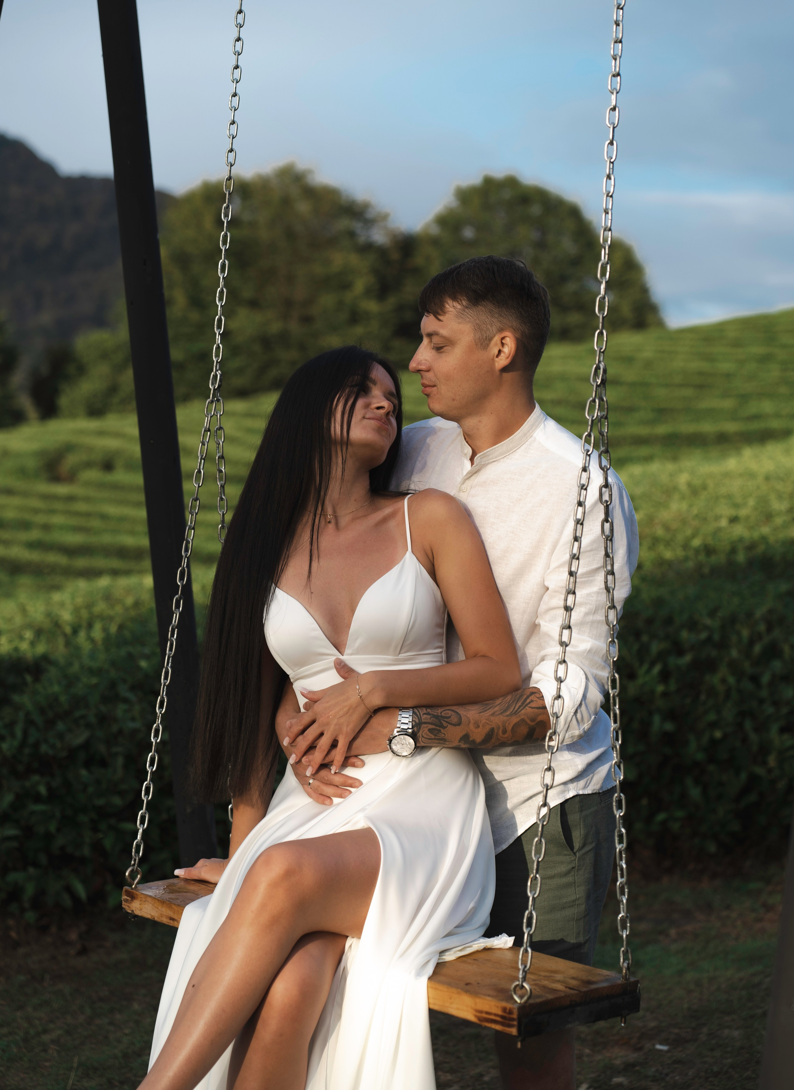 На изображении изображена пара, сидящая на качелях. Человек слева, видимо, одет в белое платье, а человек справа - в светлую рубашку и темные брюки. Фон состоит из зелени и деревьев под ясным небом. Оба носят наручные часы, а у человека справа видна татуировка на левой руке. Качели крепятся к металлической конструкции.