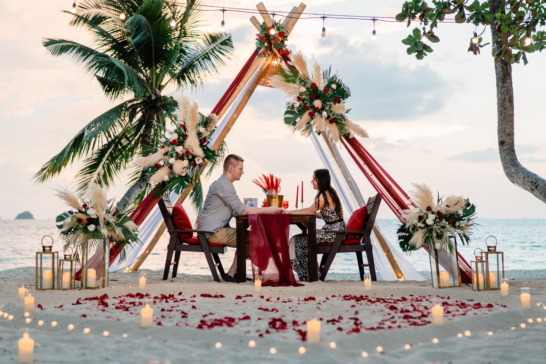 Romantic beach wedding setup on Koh Samui with a triangular floral arch, red and white draping, rose petals, candles, and ocean views.