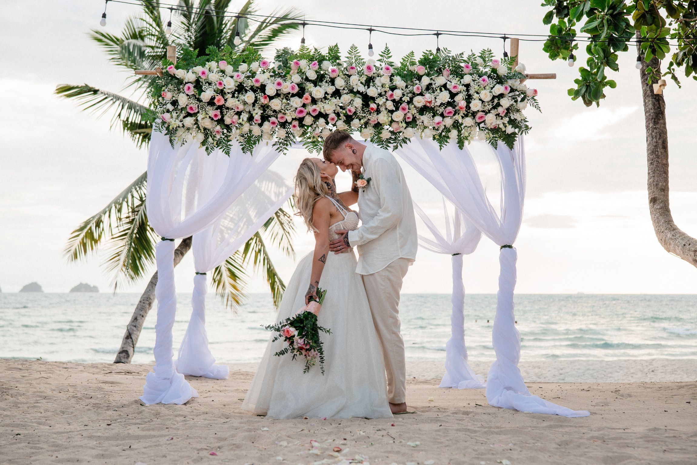 Bride and groom beneath a white-draped floral wedding arch decorated with pink flowers and tropical greenery on Koh Samui beach.