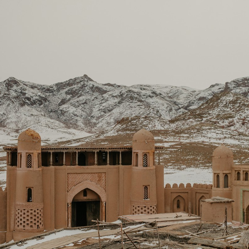 Высокий скалистый утес в пустынном ландшафте возвышается под ярким голубым небом с небольшими облаками. Поверхность скалы изрезана эрозией, придавая ей причудливую форму. Вокруг видна сухая земля с небольшими участками белого налета, возможно, из-за снега или соли. На заднем плане — более низкие скальные образования Чарынского каньона Казахстана.