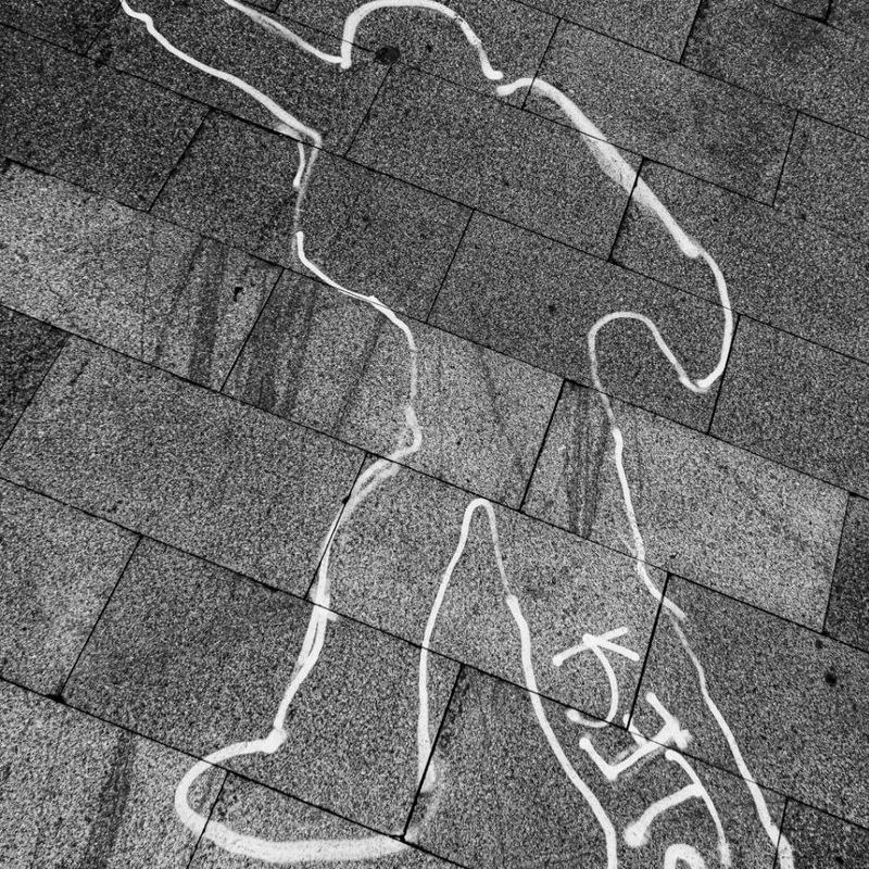 Черно-белая фотография меловой стигматы: контур лежащего человека с надписью KILLS на серой тротуарной плитке. Проект "Манифест теней" Вадима Топорёнка. Городская археология душ в дневнике фотографа-урбаниста. Урбанистическая фотография, городская философия, тени памяти.