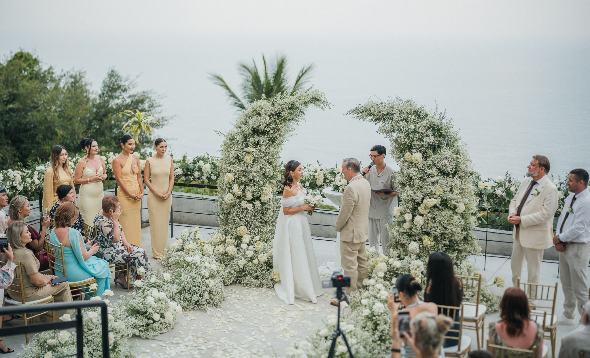 Romantic beachfront wedding ceremony in Koh Samui, Thailand, with a couple exchanging vows beneath a white floral arch surrounded by tropical greenery and guests.