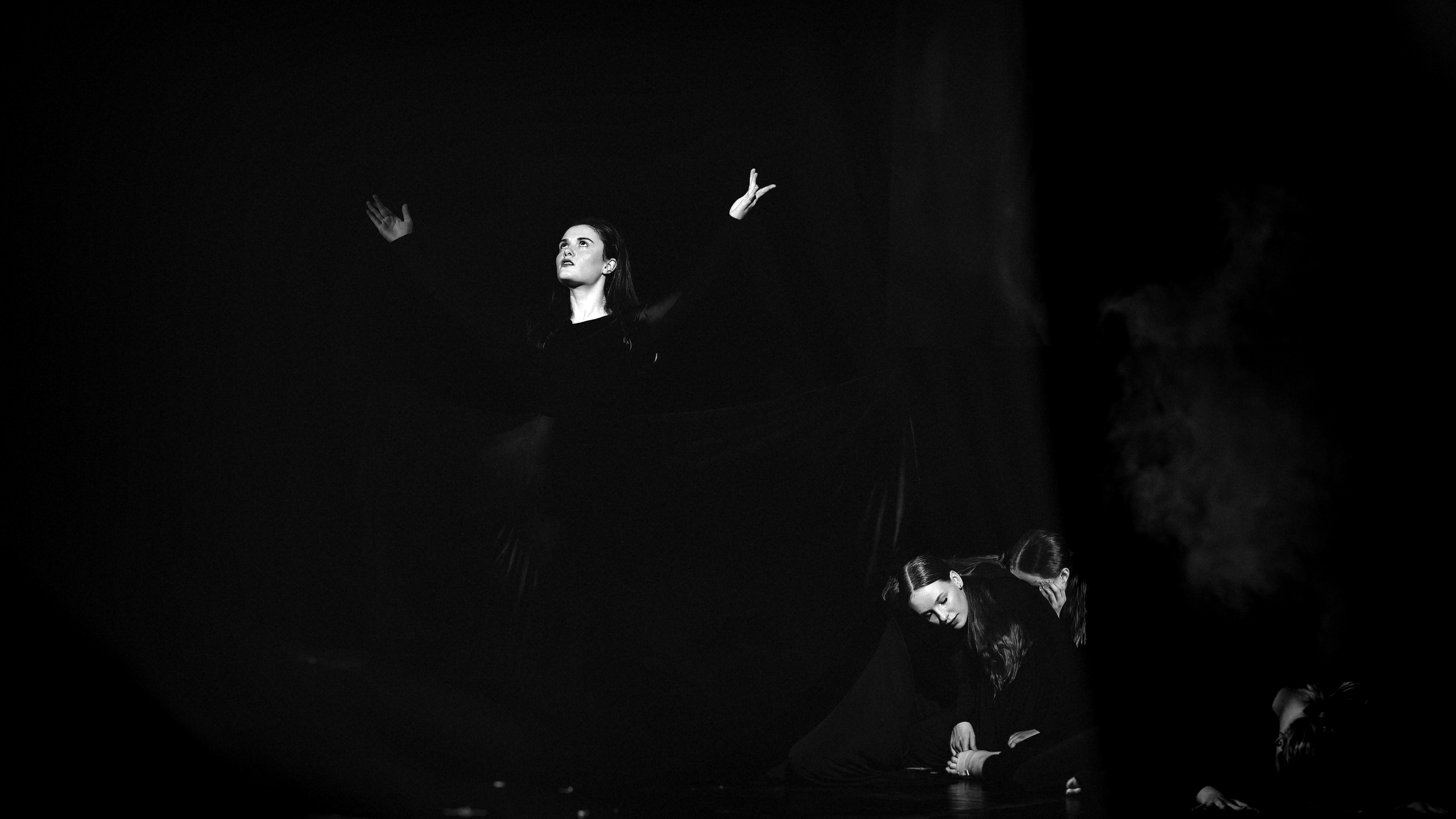 Танцоры на темной сцене с поднятыми руками и дымом, контрастное освещение, одна фигура стоит, другие на полу