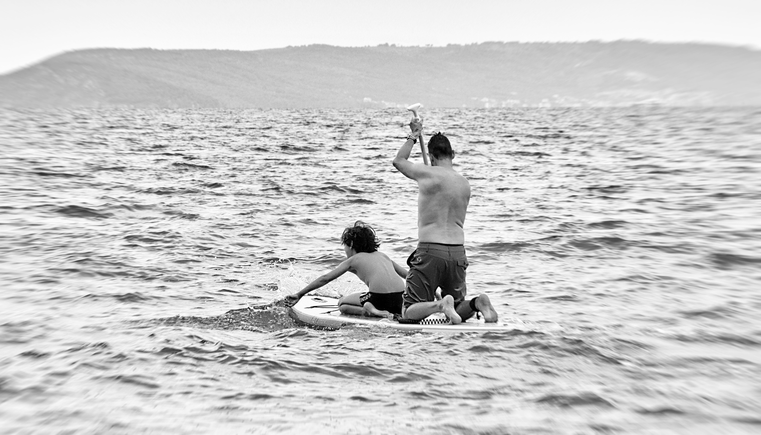 Черно-белое фото: мужчина и ребенок на сап-борде в море, взрослый стоит и гребет веслом, ребенок сидит на доске, на горизонте горы, документальная семейная фотография
