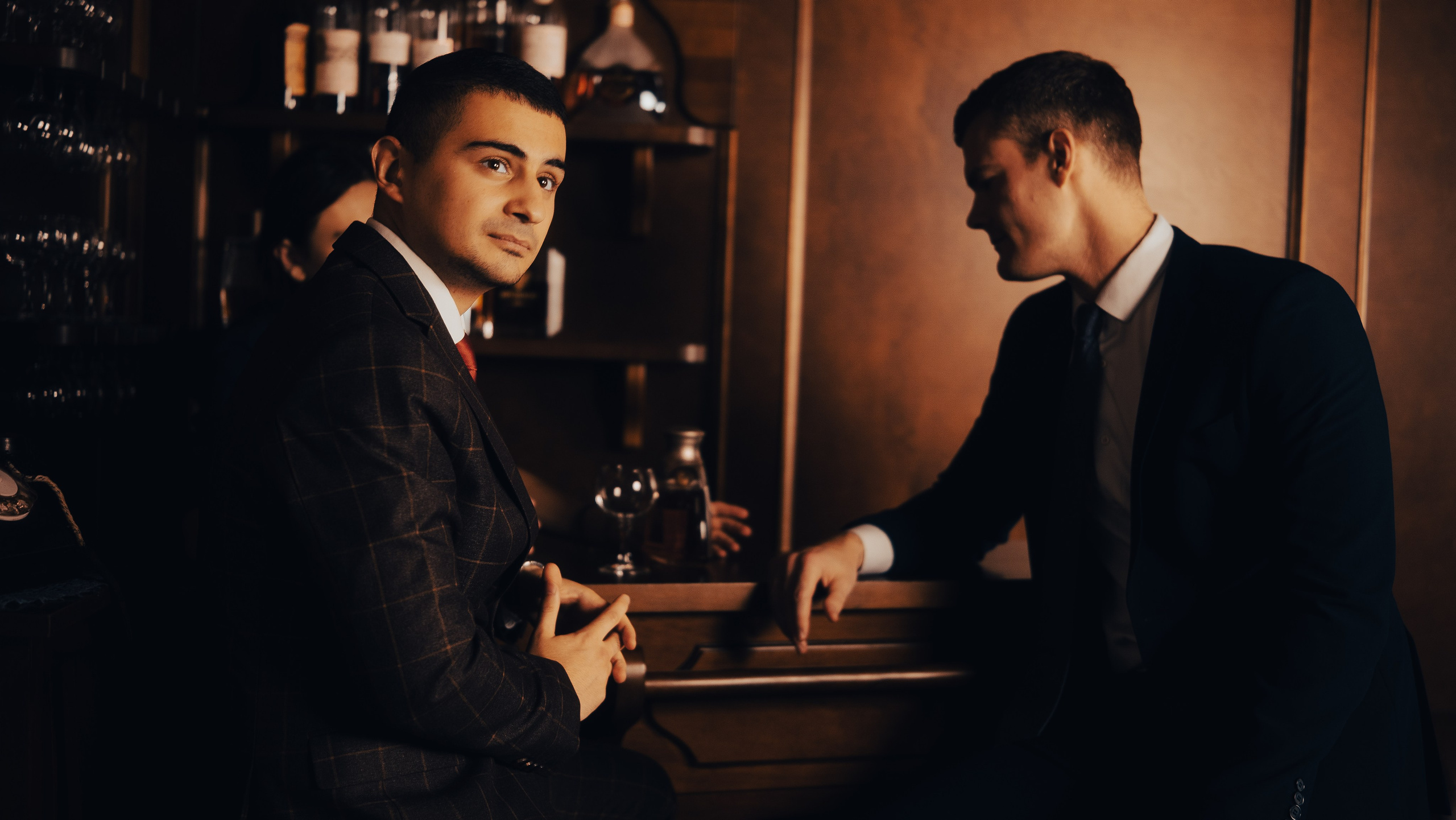 На фотографии изображены два мужчины в строгих костюмах, находящиеся в интерьере, напоминающем бар или частный клуб. Атмосфера снимка выдержана в тёплых, приглушённых тонах, создающих ощущение уюта и лёгкой загадочности. Мужчина слева: сидит, слегка повернувшись к камере; смотрит прямо в объектив с лёгкой полуулыбкой, выражение лица уверенное, но не вызывающее; одет в тёмный клетчатый пиджак, белую рубашку и красный галстук; руки сложены вместе, поза расслабленная, но собранная; его образ производит впечатление человека, привыкшего контролировать ситуацию. Мужчина справа: стоит, слегка наклонившись вперёд, опираясь рукой на барную стойку; взгляд направлен на собеседника (мужчину слева), выражение лица серьёзное, сосредоточенное; одет в классический тёмный костюм, белую рубашку и тёмный галстук; поза динамичная, создаёт ощущение вовлечённости в разговор или переговоры. Фон и детали: за спинами мужчин видны полки с бутылками алкоголя, что подчёркивает барную атмосферу; на стойке стоят бокалы и бутылка, добавляя реалистичности сцене; освещение мягкое, с тёплым оттенком, акцентирует внимание на лицах и костюмах героев; интерьер выполнен в классическом стиле с элементами дерева и тёмных оттенков. Общее настроение снимка: Фотография передаёт атмосферу серьёзного, но не напряжённого диалога между двумя мужчинами. Композиция, освещение и позы героев создают ощущение драмы с элементами элегантности и стиля. Снимок может ассоциироваться с сценами из кино о деловых переговорах или дружеской беседе в приватной обстановке.