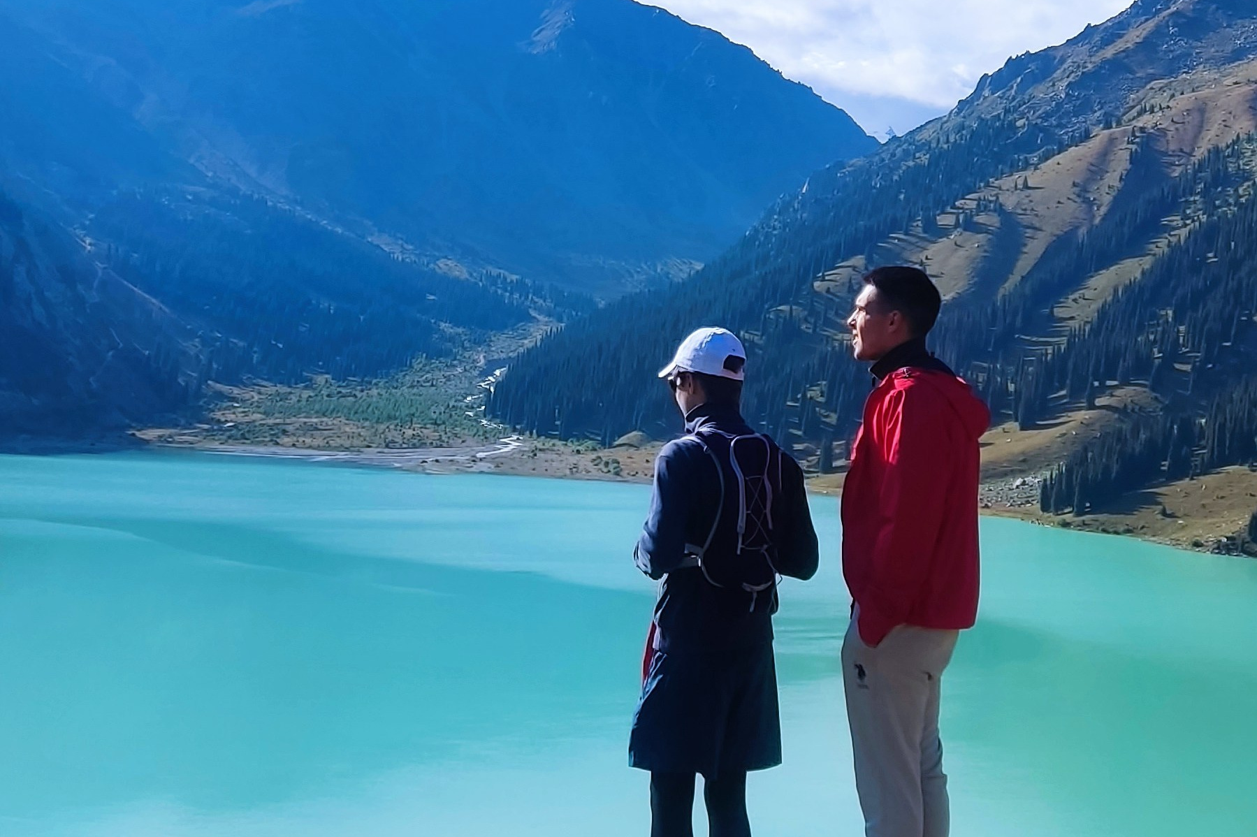 Горное озеро Иссык в Алматинской области, Казахстан, с бирюзовой водой на фоне осеннего леса с яркими золотистыми деревьями, окружённого высокими горами, покрытыми хвойным лесом. На горизонте видны заснеженные вершины под облачным небом