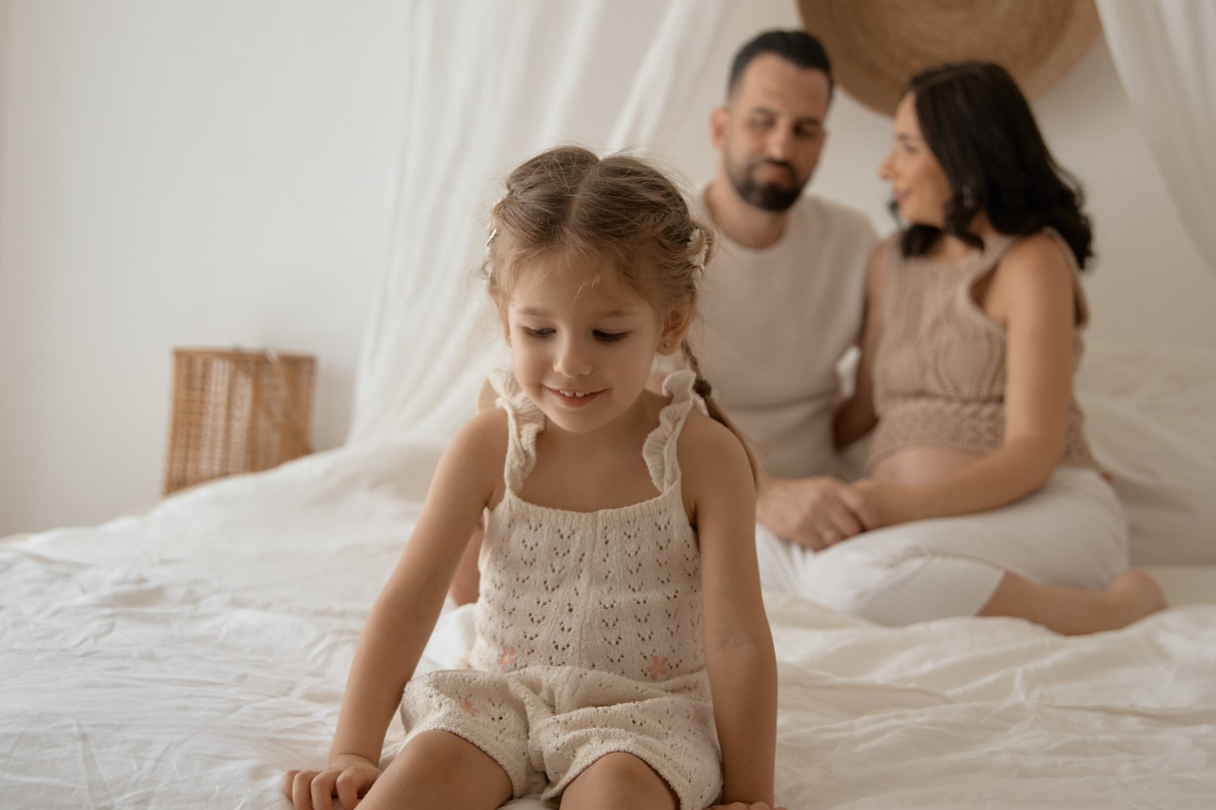 A young girl sits on a bed with her parents in the background, sharing a tender moment together
