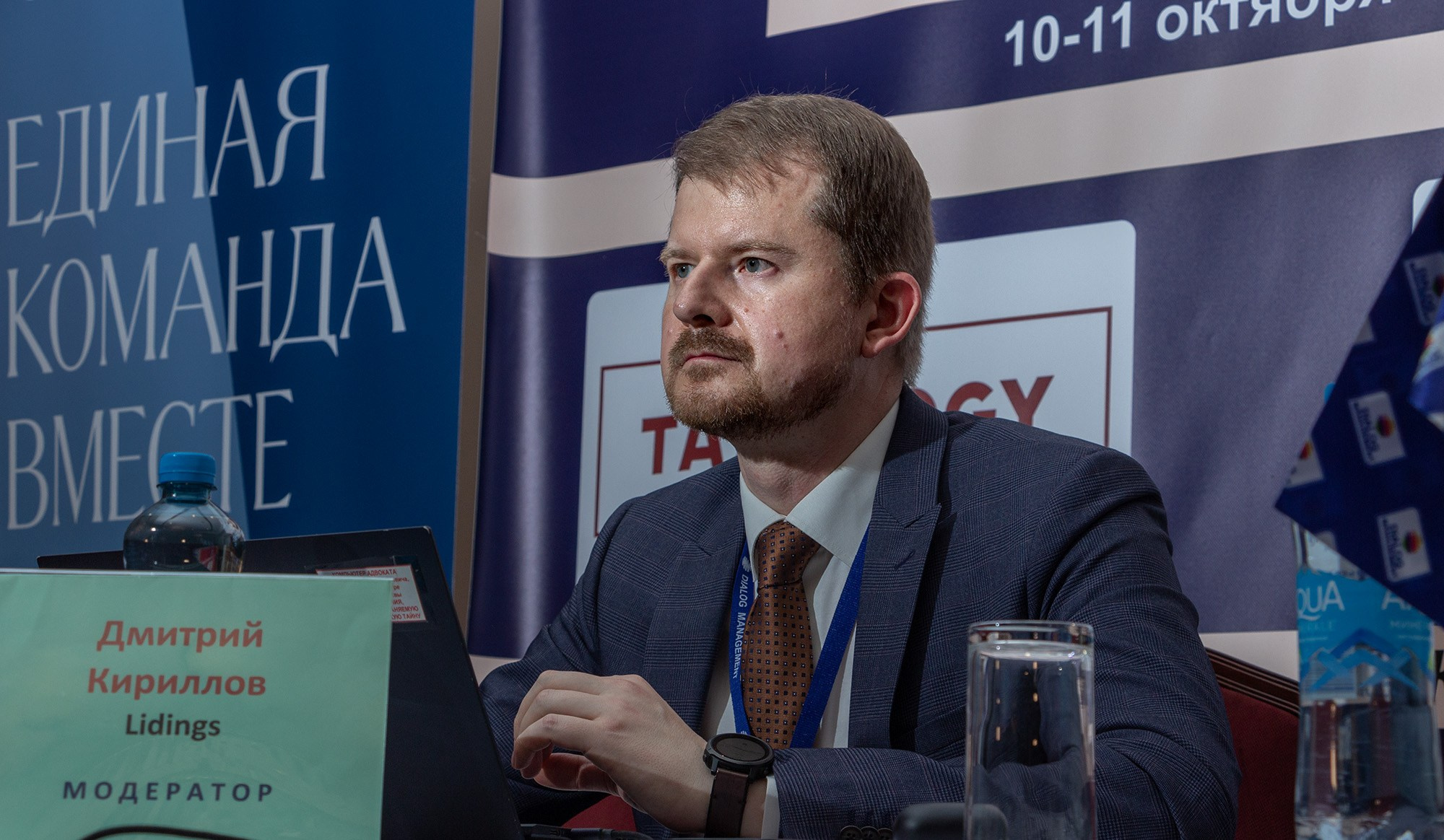 Модератор за столом с именной табличкой на сцене деловой конференции