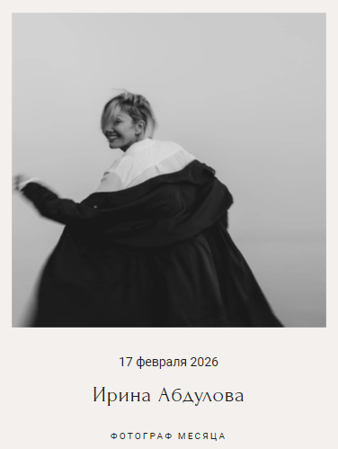 Девушка в светлой блузе и тёмном пальто улыбается, будто кружится. Фон нейтральный, атмосфера лёгкая, игривая.