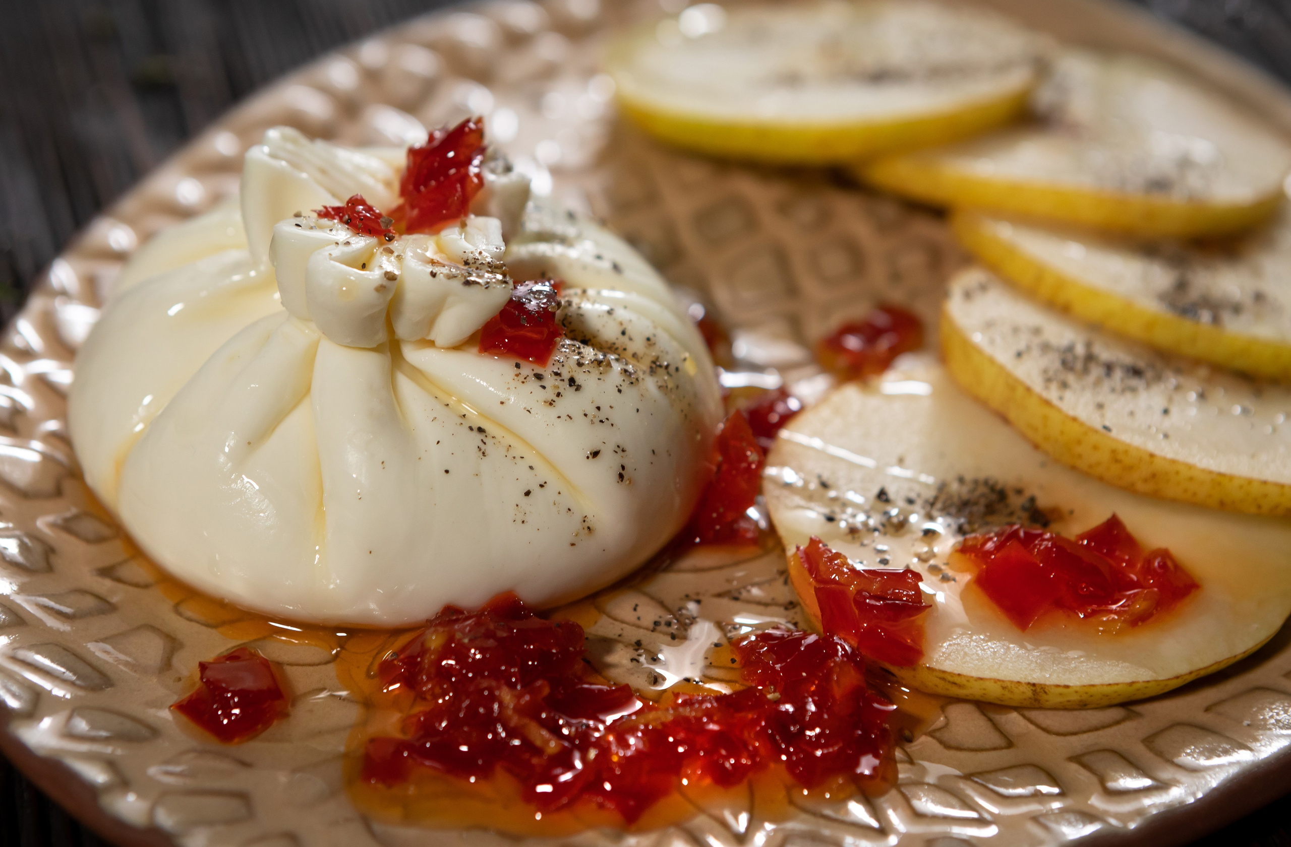 Мешочек сыра буррата с хвостиком на белой прямоугольной тарелке со специями, помидорками черри и красным салатом