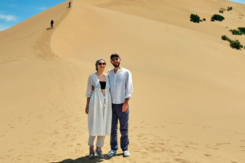 Человек позирует на фоне бирюзового озера Иссык в районе Алматы и гор. Он стоит на большом камне, одет в ярко-зеленую толстовку, черные штаны и светлую обувь, а также оранжевую панаму. В руках держит очки. За ним виднеются деревья, лесистые склоны и горные вершины под облачным небом. Атмосфера спокойная и природная.