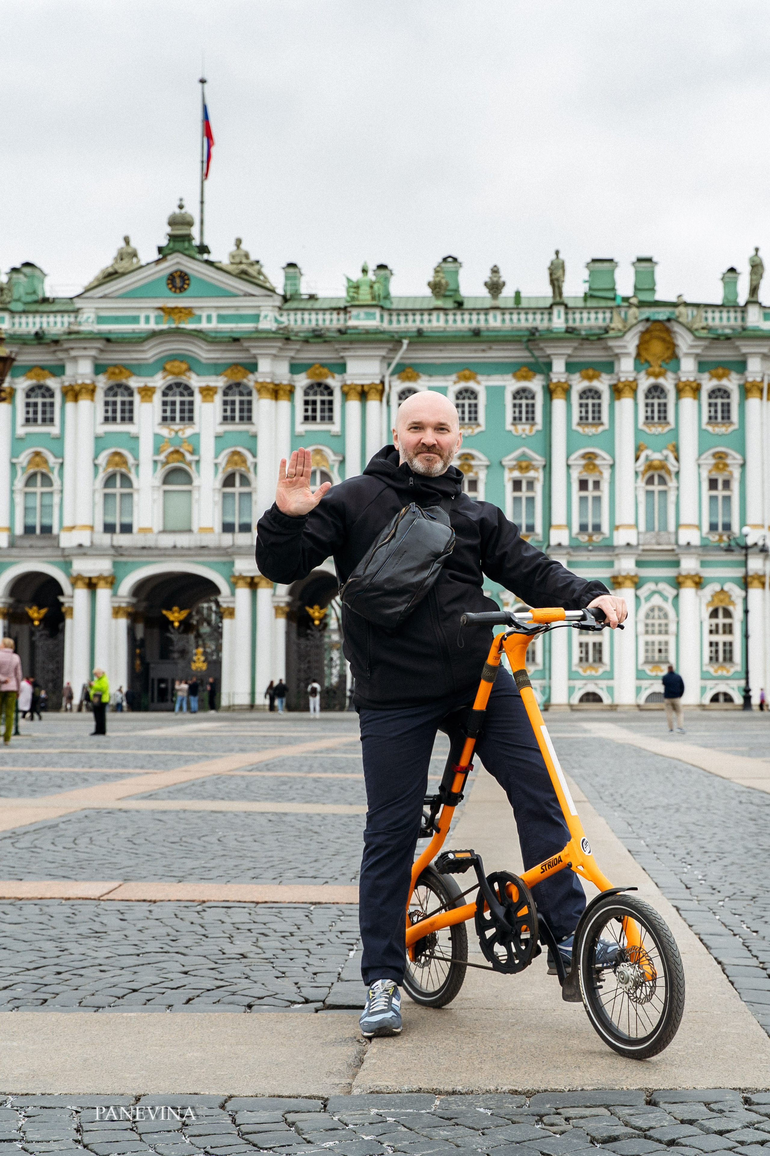 Счастливый экскурсовод машет Рукой стоя на дворцовой площади за спиной зимний дворец