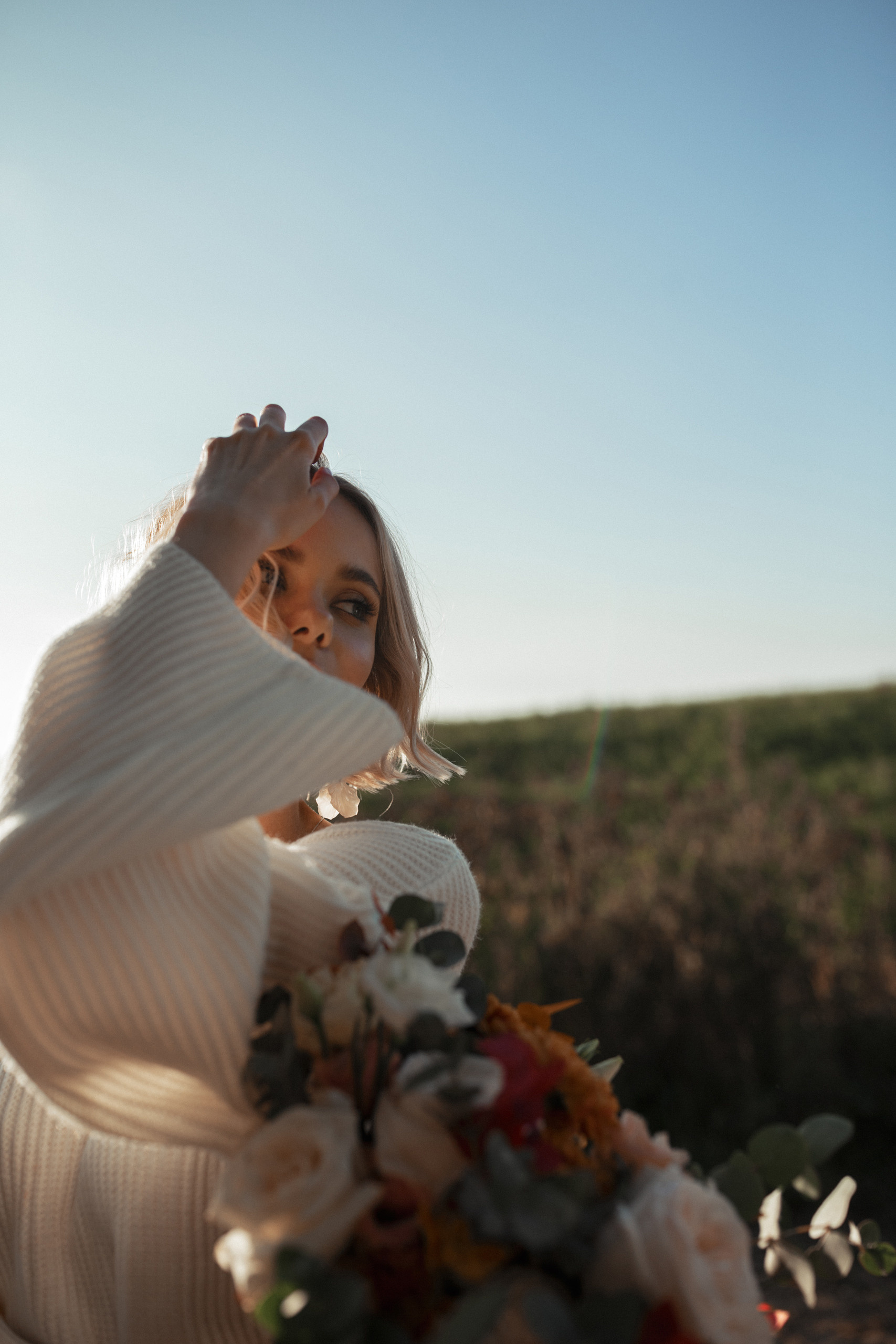 Невеста в белом платье с большими руками поправляет свою прическу, глядя на жениха.