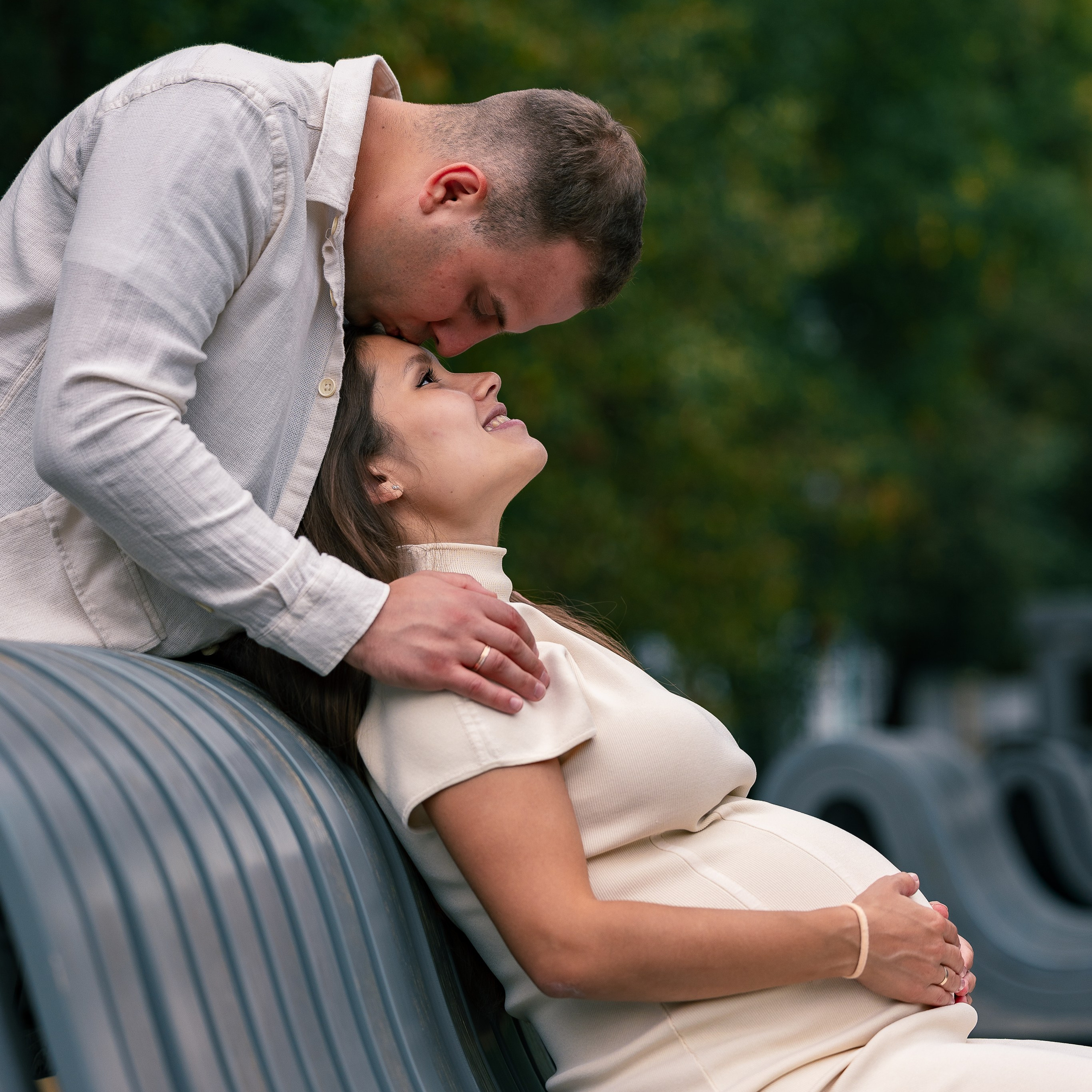 Фотография беременной девушки, которая сидит на скамье, а ее супруг стоит сзади и целует в лоб