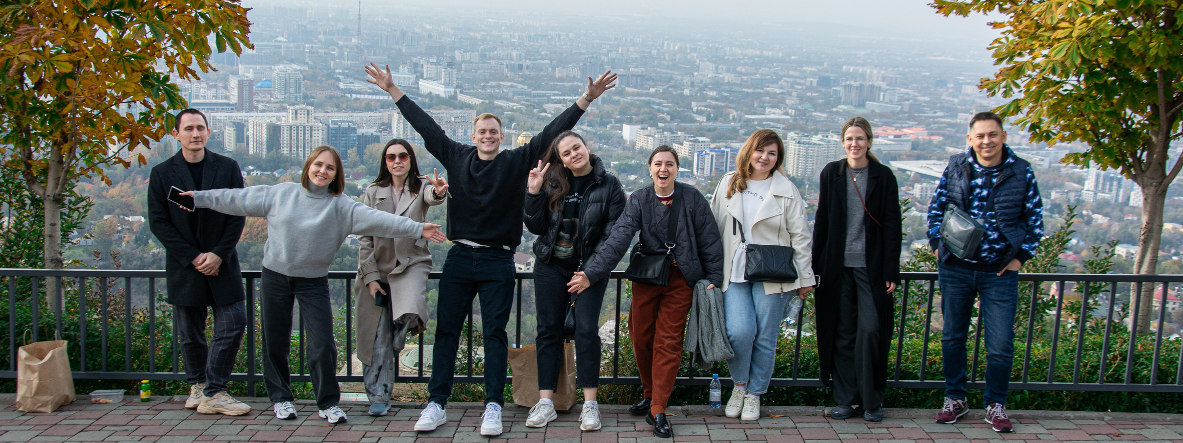 Группа из шести человек в зимней одежде прыгает на фоне заснеженных гор. Все выглядят счастливыми и веселыми, наслаждаясь снежной атмосферой и солнечным днем. Вдали виднеются горные склоны с густыми лесами, а также канатная дорога Шымбулак. Небо ясное с небольшими облаками, создавая живописный зимний пейзаж.