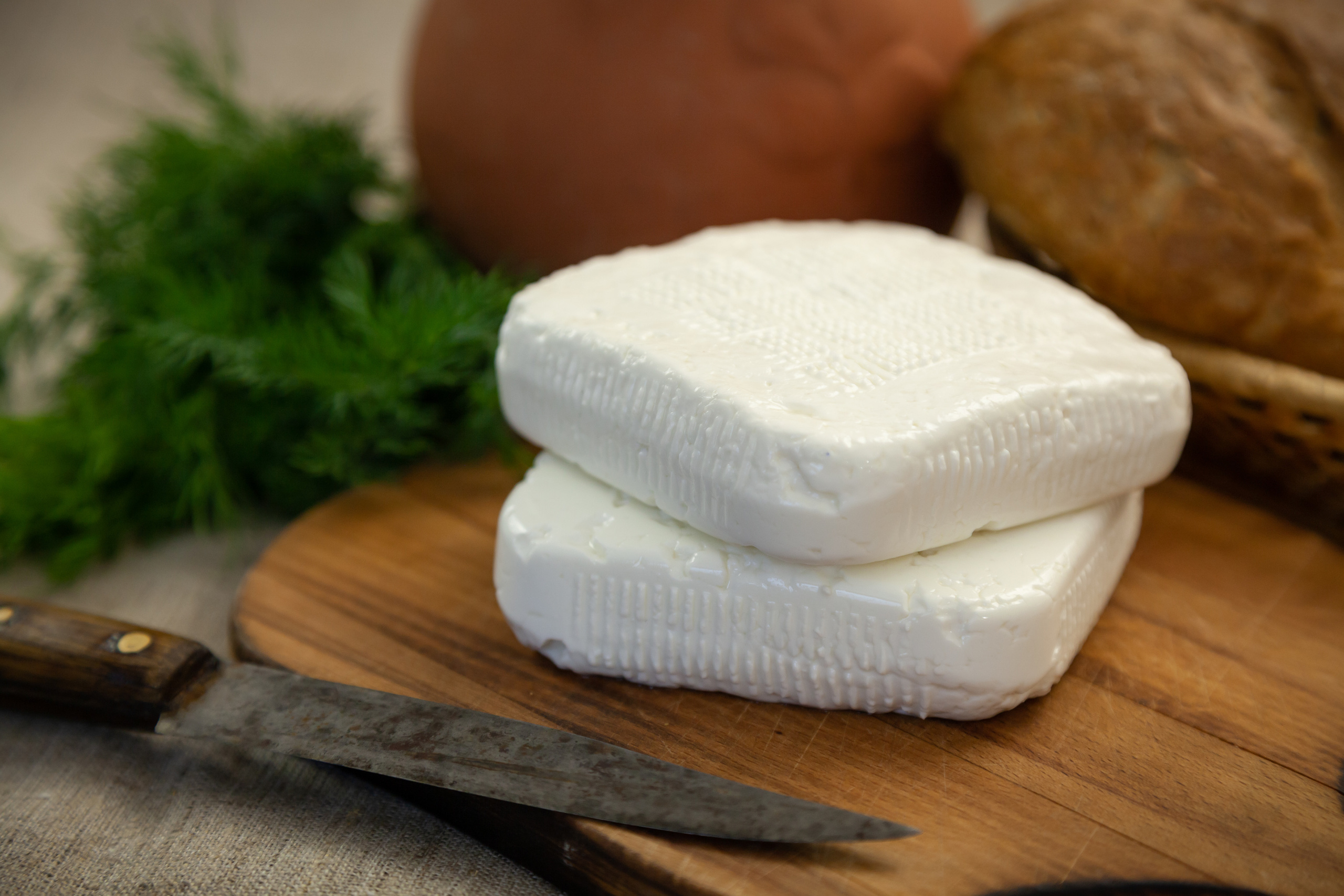 Два куска имеретинского сыра на грубой разделочной доске. Рядом старый нож, на фоне зелень, кувшин и хлеб.