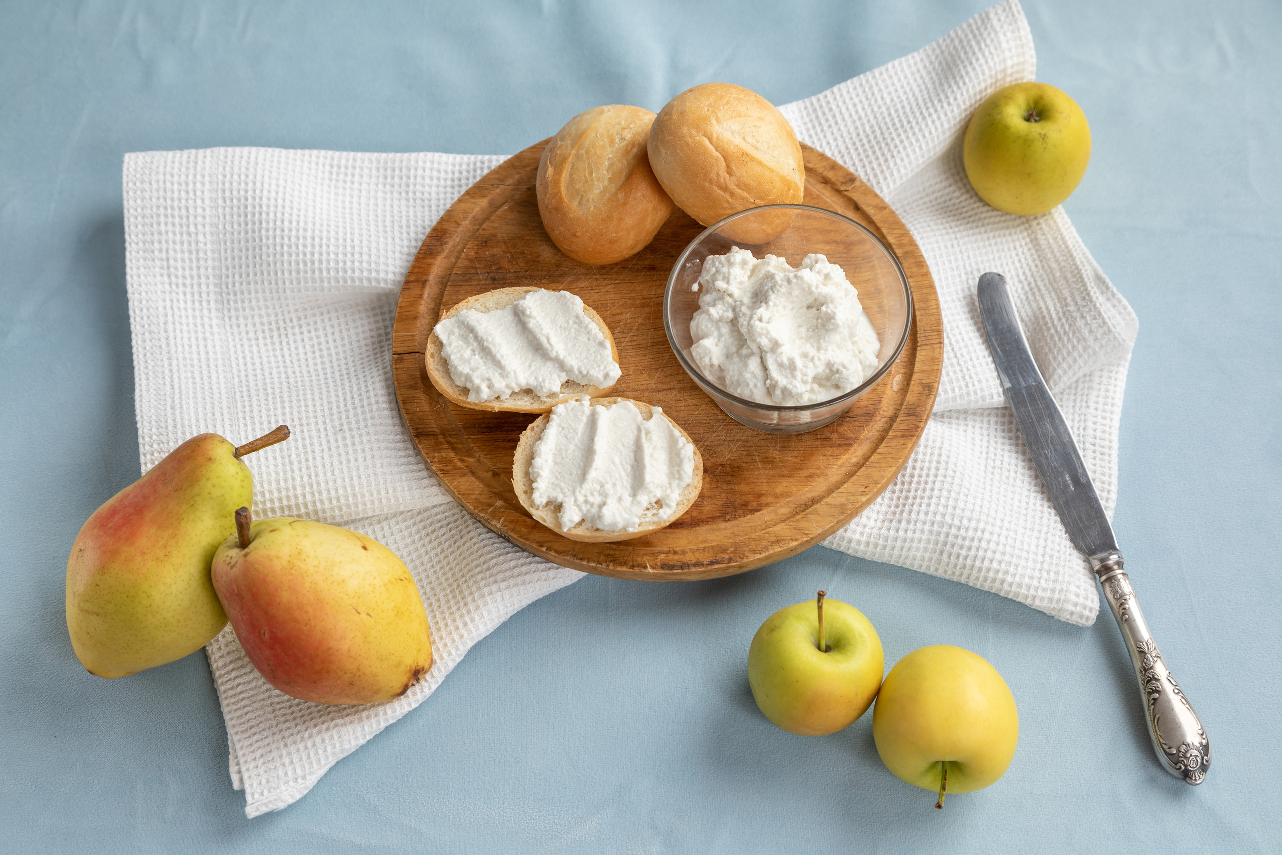 Натюрморт из бутербродов с сыром рикотта и булочками на деревянной круглой досочке с грушами и яблоками, все это на белом полотенце и на голубой скатерти.