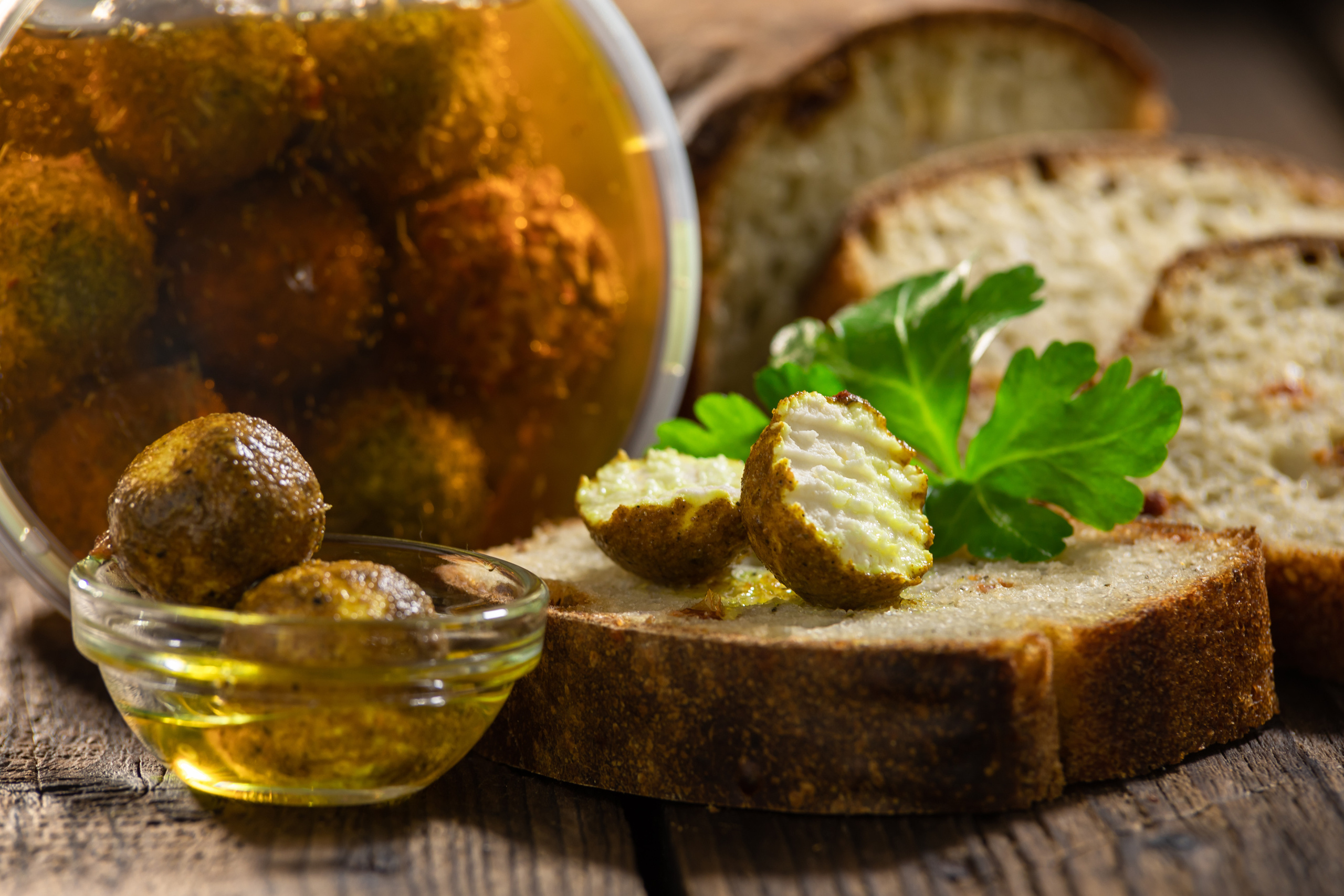 Сырные шарики в масле крупным планом в маленькой стеклянной чашке и на куске хлеба на фоне банки с сырными шариками и буханкой хлеба