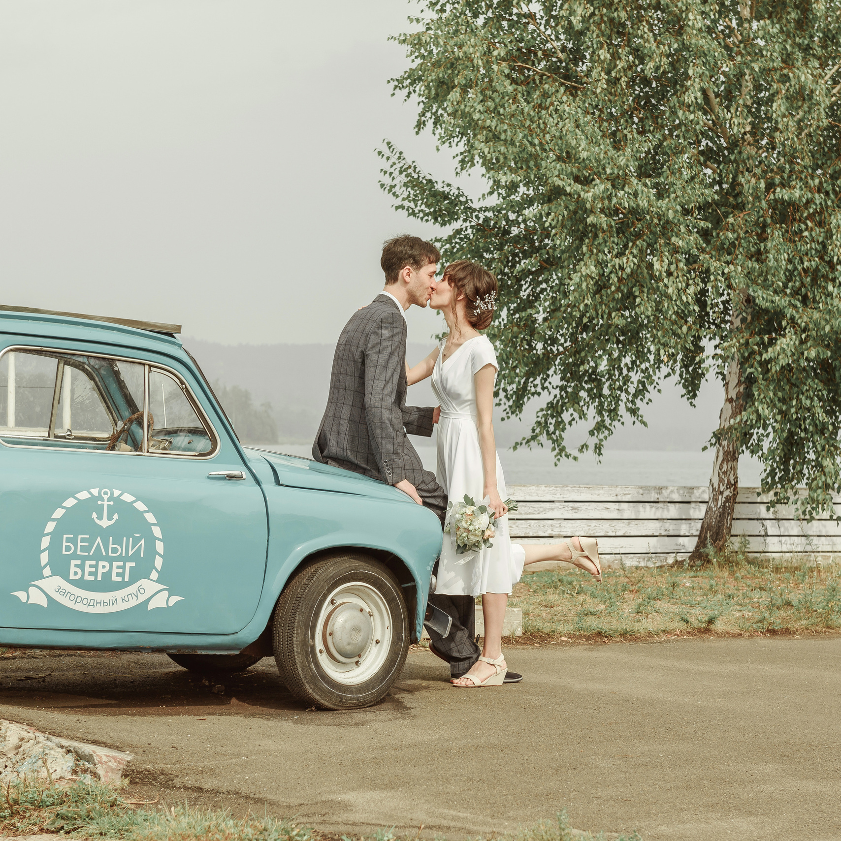 Парень и девушка стоят возле голубой машины в день свадьбы и целуются