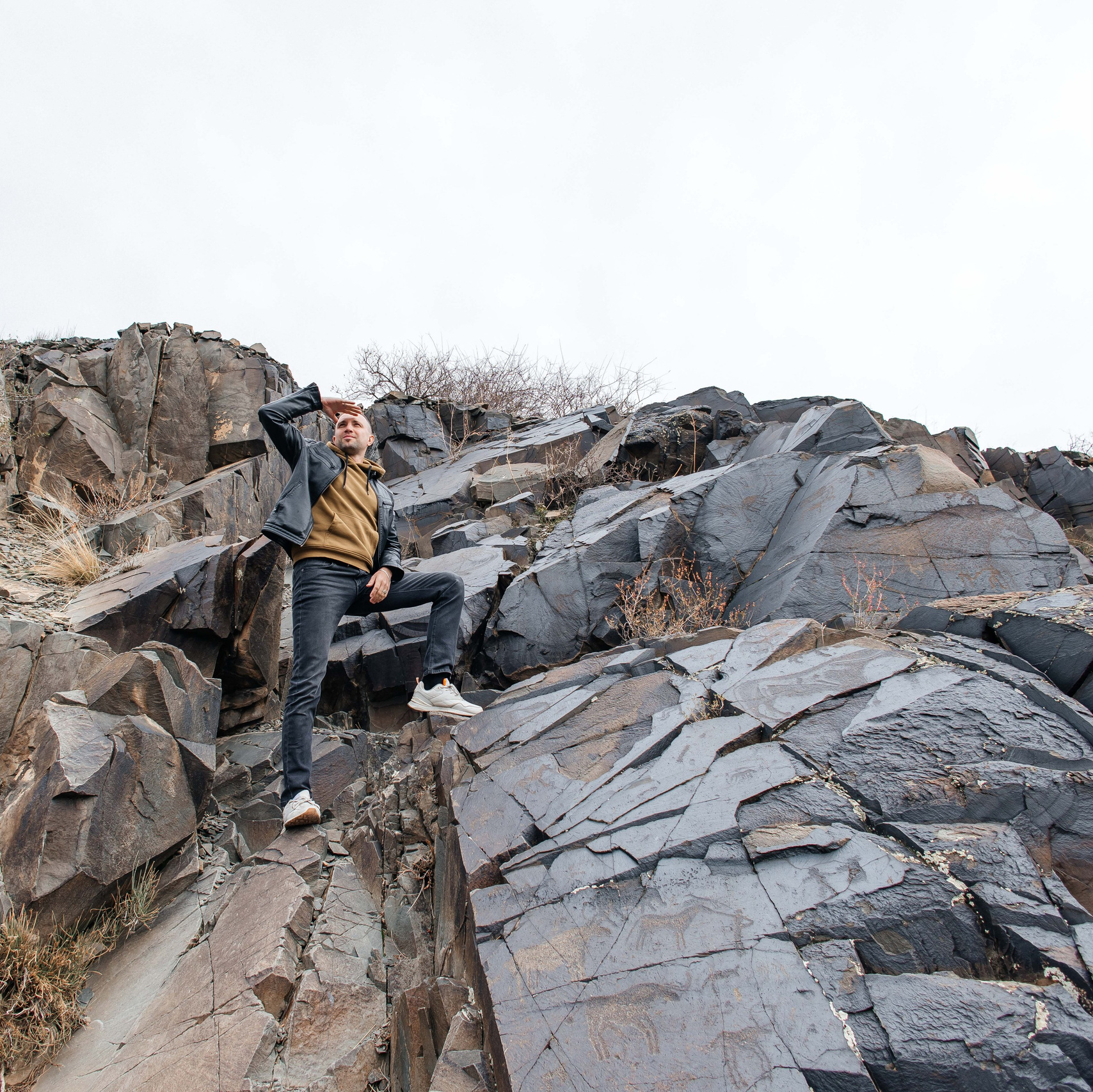 Высокий скалистый утес в пустынном ландшафте возвышается под ярким голубым небом с небольшими облаками. Поверхность скалы изрезана эрозией, придавая ей причудливую форму. Вокруг видна сухая земля с небольшими участками белого налета, возможно, из-за снега или соли. На заднем плане — более низкие скальные образования Чарынского каньона Казахстана.