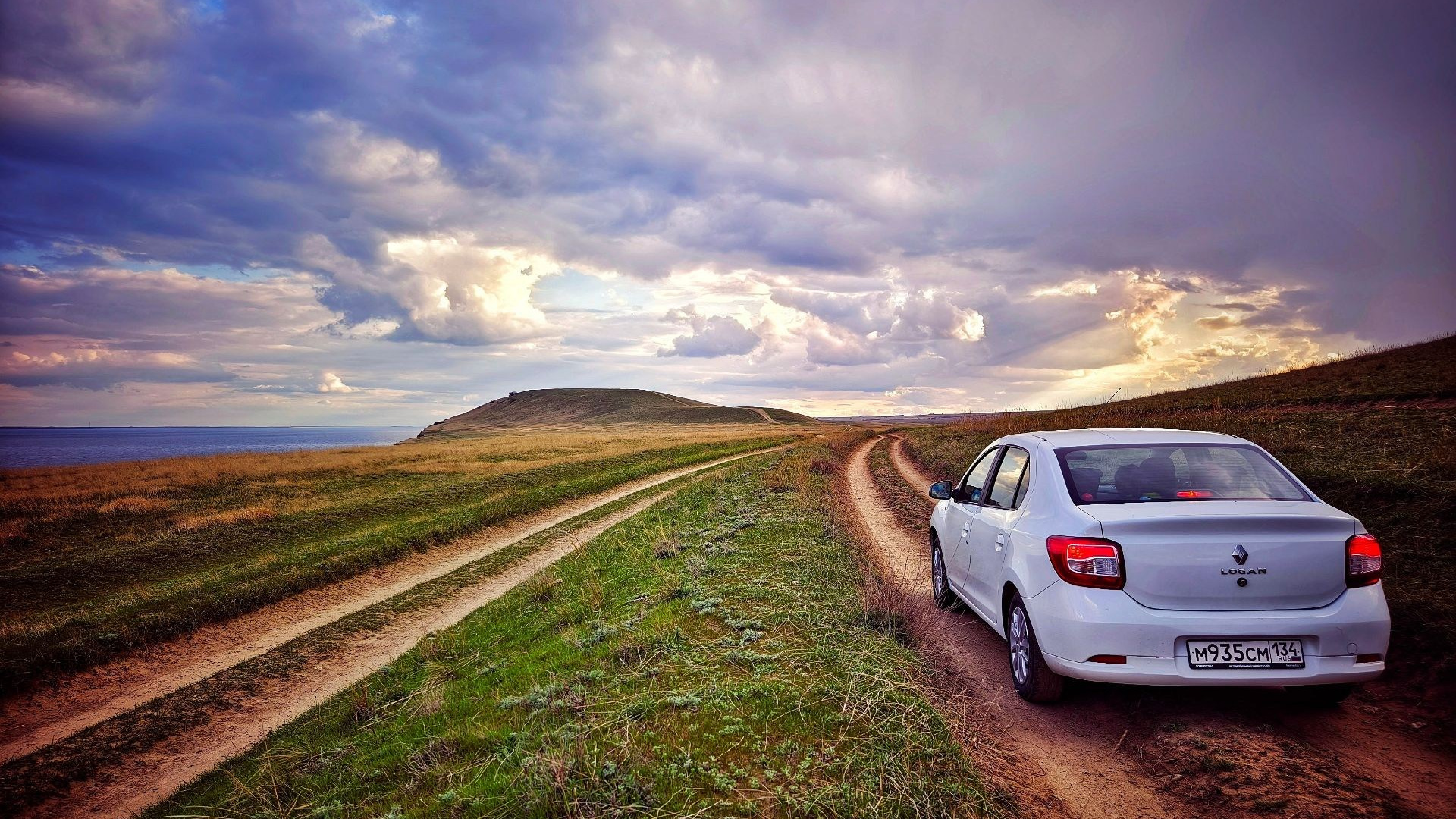 Белый Renault Logan на грунтовой дороге у моря, закат и облака создают атмосферу спокойствия