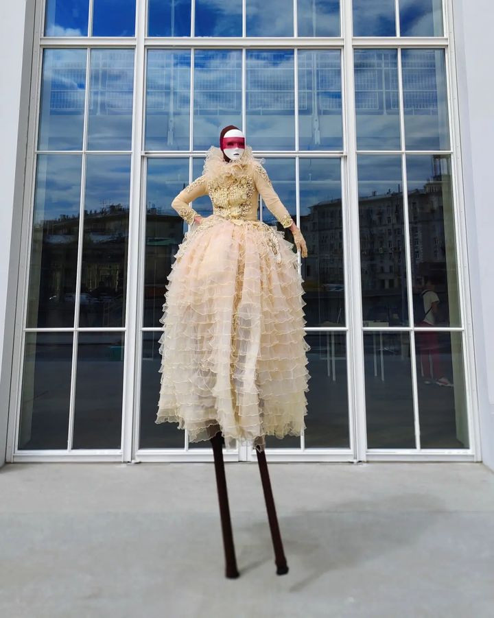 Ходулист в бежевом платье встречает гостей на велком-зоне у ресторана.