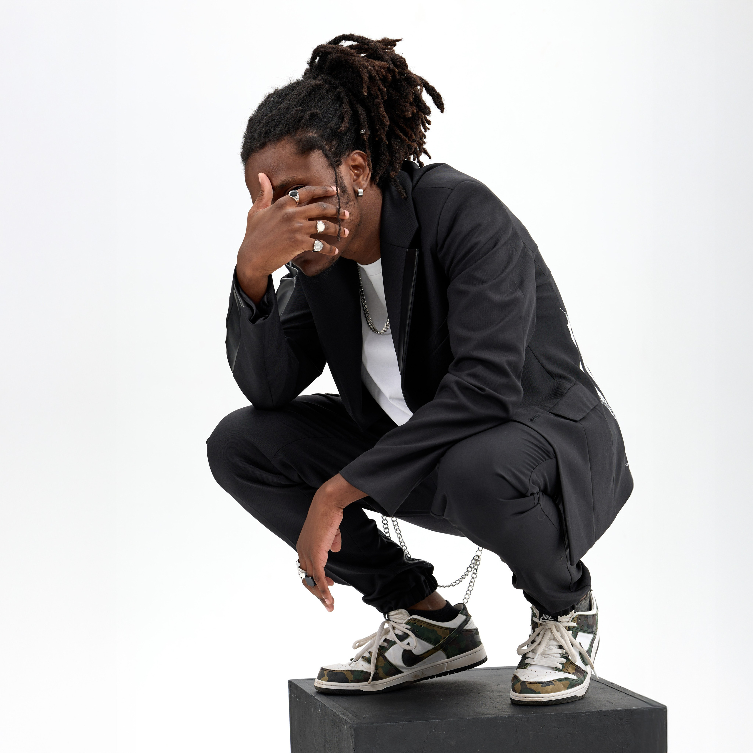 Съёмка каталога новой коллекции костюмов для бренда одежды: мужчина с дредами в чёрном костюме стоит в динамичной позе, присев на один колено. Он прикрывает лицо рукой, создавая стильный, задумчивый образ. На нём белая футболка и кроссовки Nike. Фон нейтральный, белый