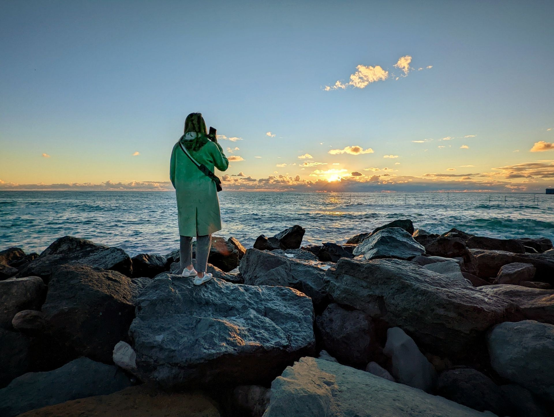 Девушка в зелёном плаще стоит на скалах у моря и любуется закатом