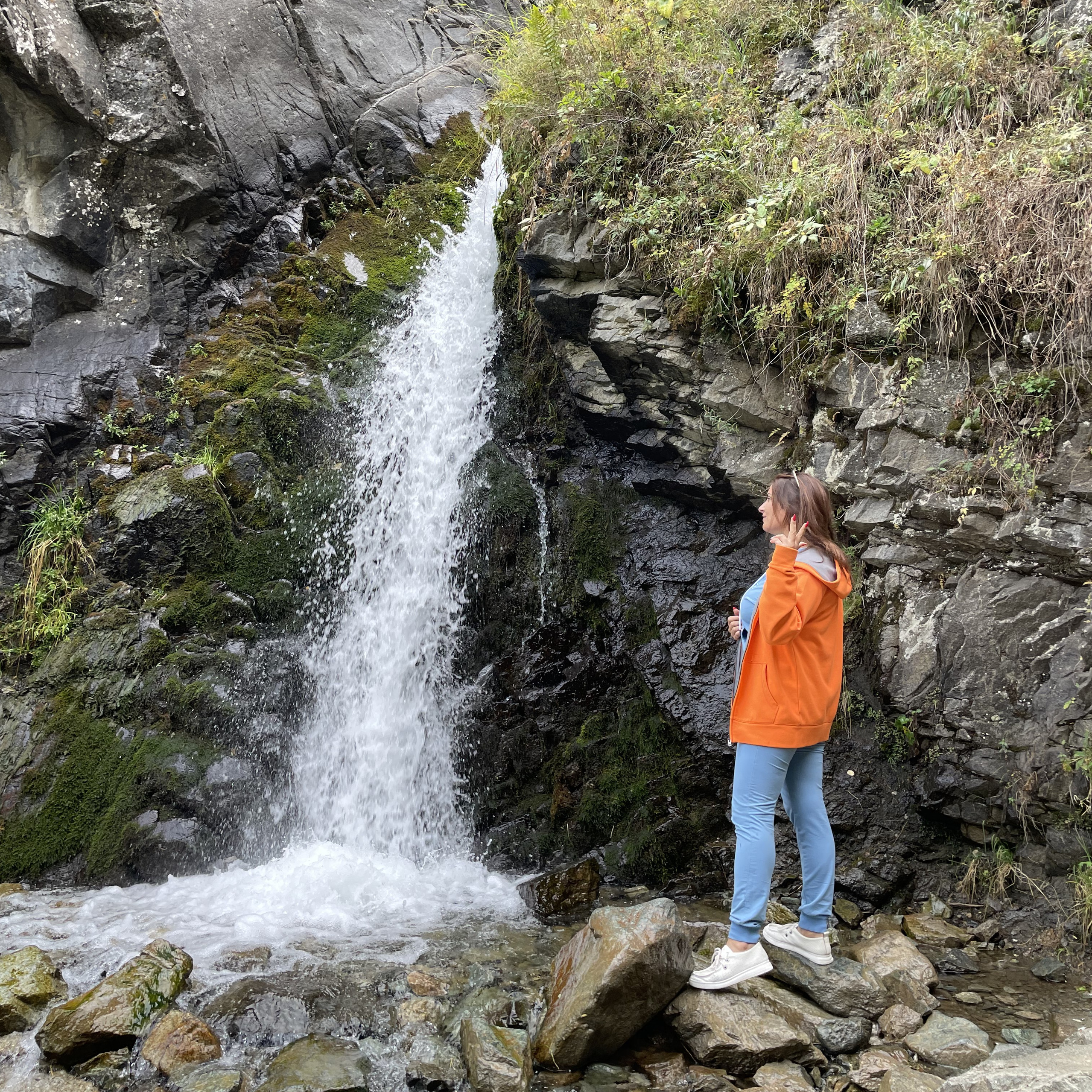 Женщина стоит рядом со скульптурой медведя на фоне красивого Медвежьего водопада в районе Алматы, спадающего по скалистым утёсам. На вершине скал и кустарниках виден свежевыпавший снег, что подчёркивает прохладную атмосферу. Каменистая местность у подножия водопада добавляет природной аутентичности сцене.