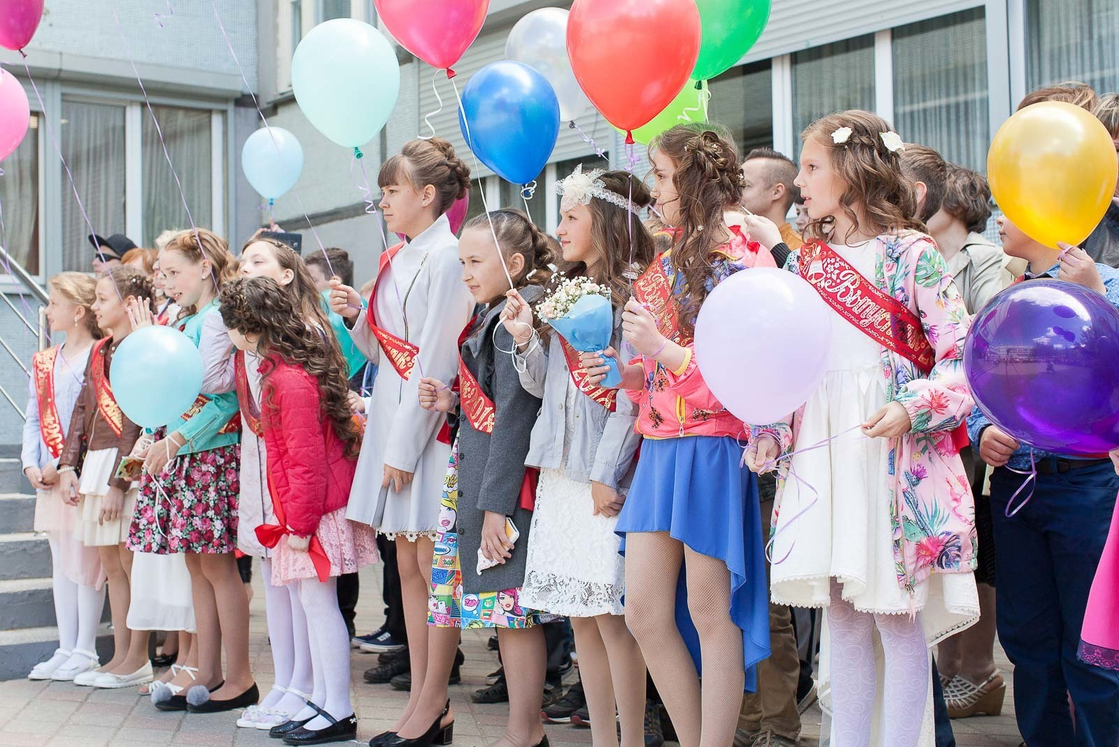 Выпускники 4 класса стоят на линейке у школы с шарами и лентами выпускника.