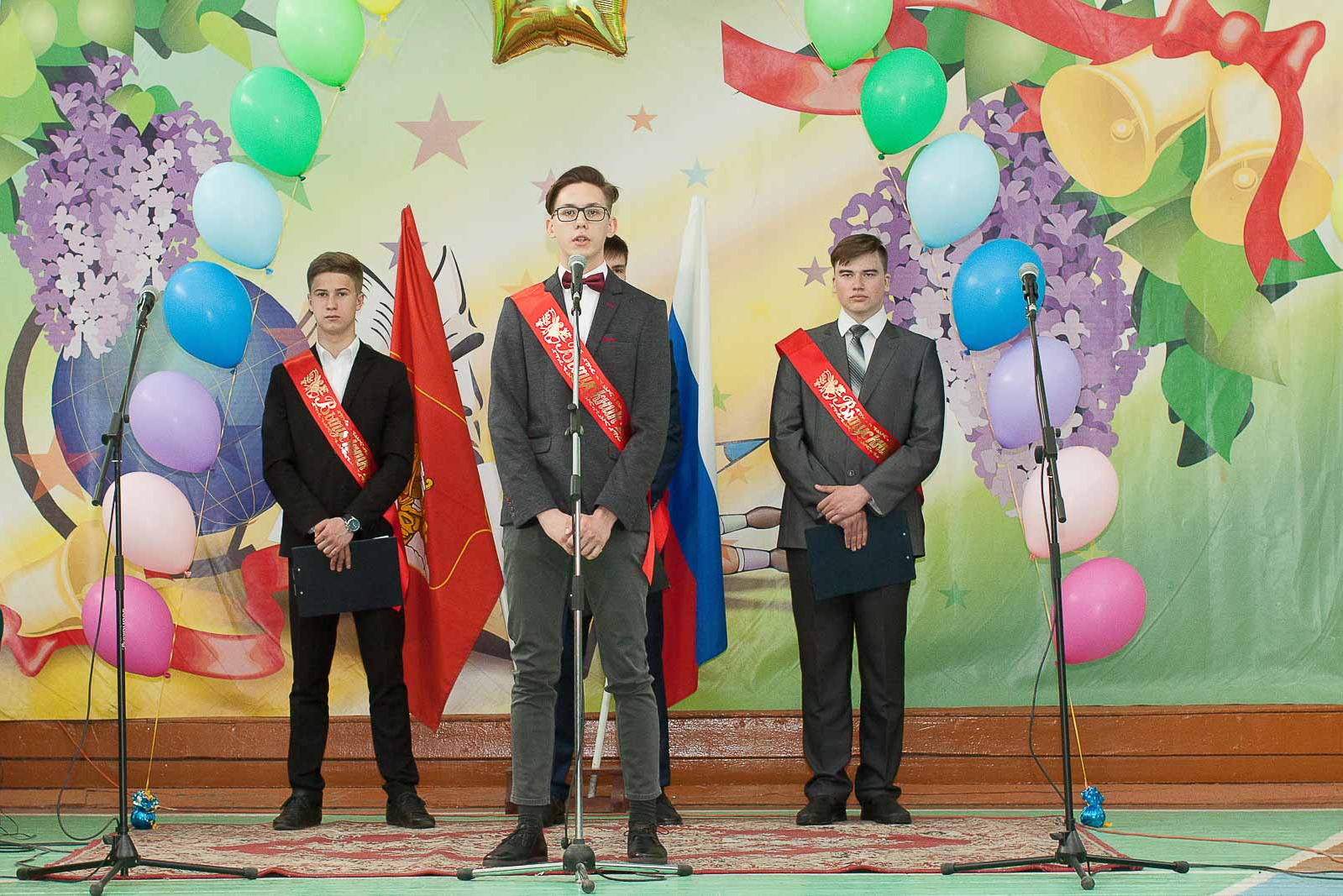 Три выпускника 11 класса с красными лентами стоят у микрофонов в школьном спортзале.