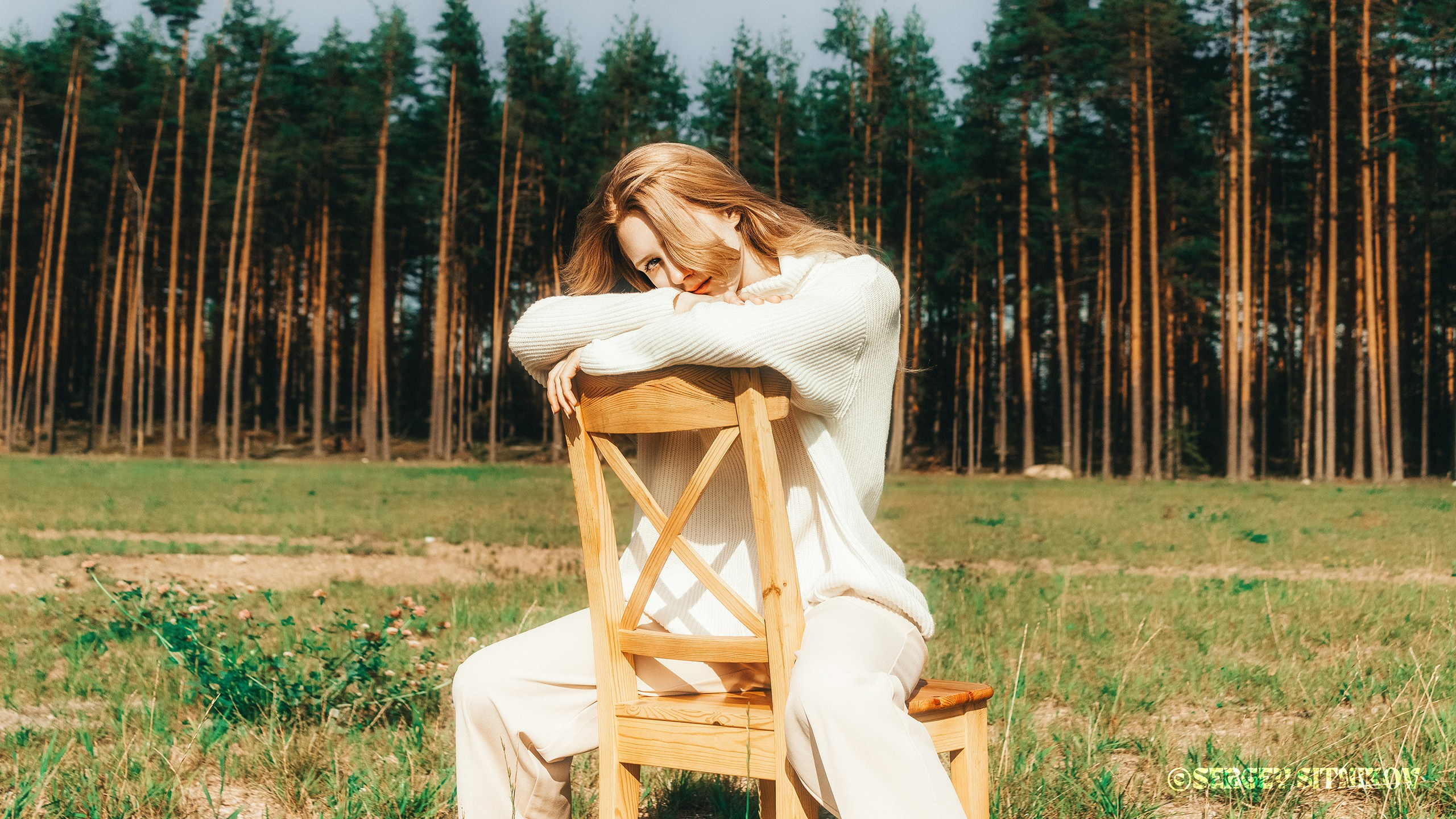 Девушка в светлой одежде сидит на деревянном стуле на поляне у соснового леса, атмосферный портрет на природе при жёстком солнечном дневном свете