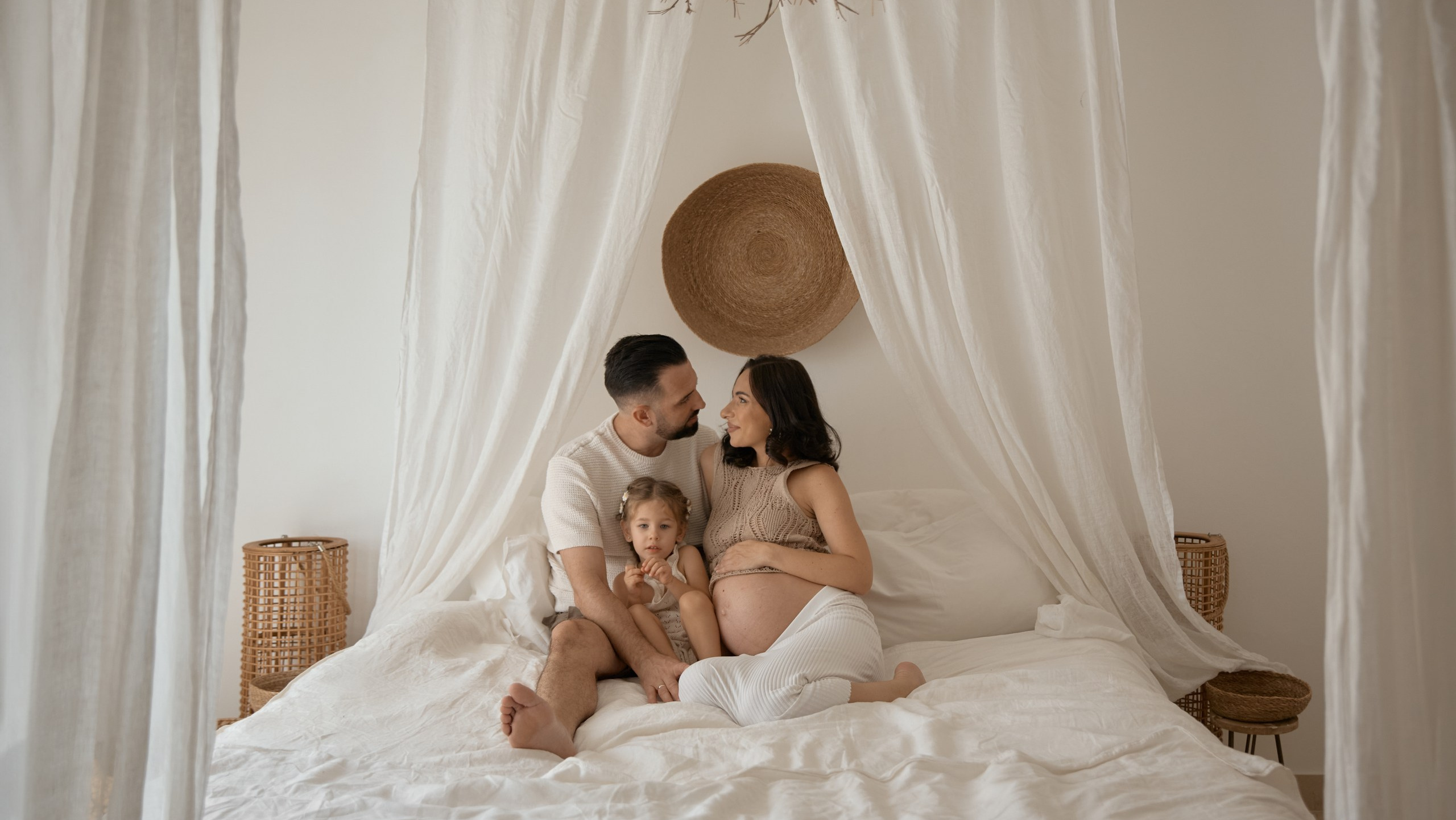 A family of three, including a pregnant mother, father, and young daughter, sits together on a bed surrounded by white curtains, sharing a loving moment