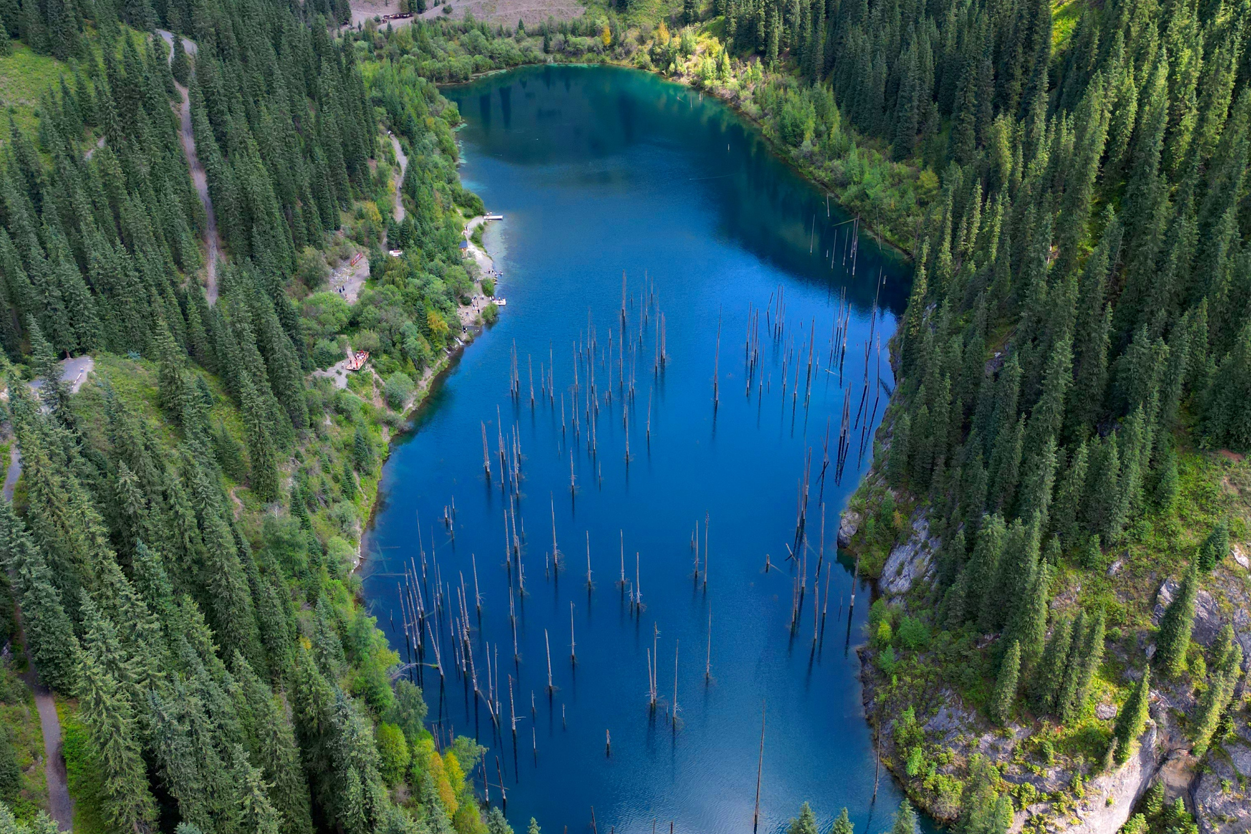 Горное озеро Иссык в Алматинской области, Казахстан, с бирюзовой водой на фоне осеннего леса с яркими золотистыми деревьями, окружённого высокими горами, покрытыми хвойным лесом. На горизонте видны заснеженные вершины под облачным небом