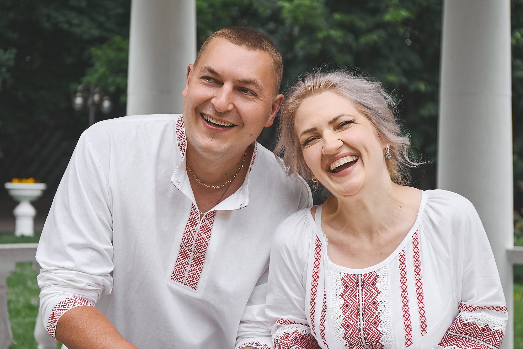 Улыбающаяся пара в традиционных русских вышиванках позирует на фоне белой колоннады в парке. русская вышиванка, традиционная одежда, семейная фотосессия, пара в вышиванках, этностиль, русская культура, национальные наряды, фотосессия в парке, семейная любовь, народный орнамент, русскиетрадиции, вышиванка фото