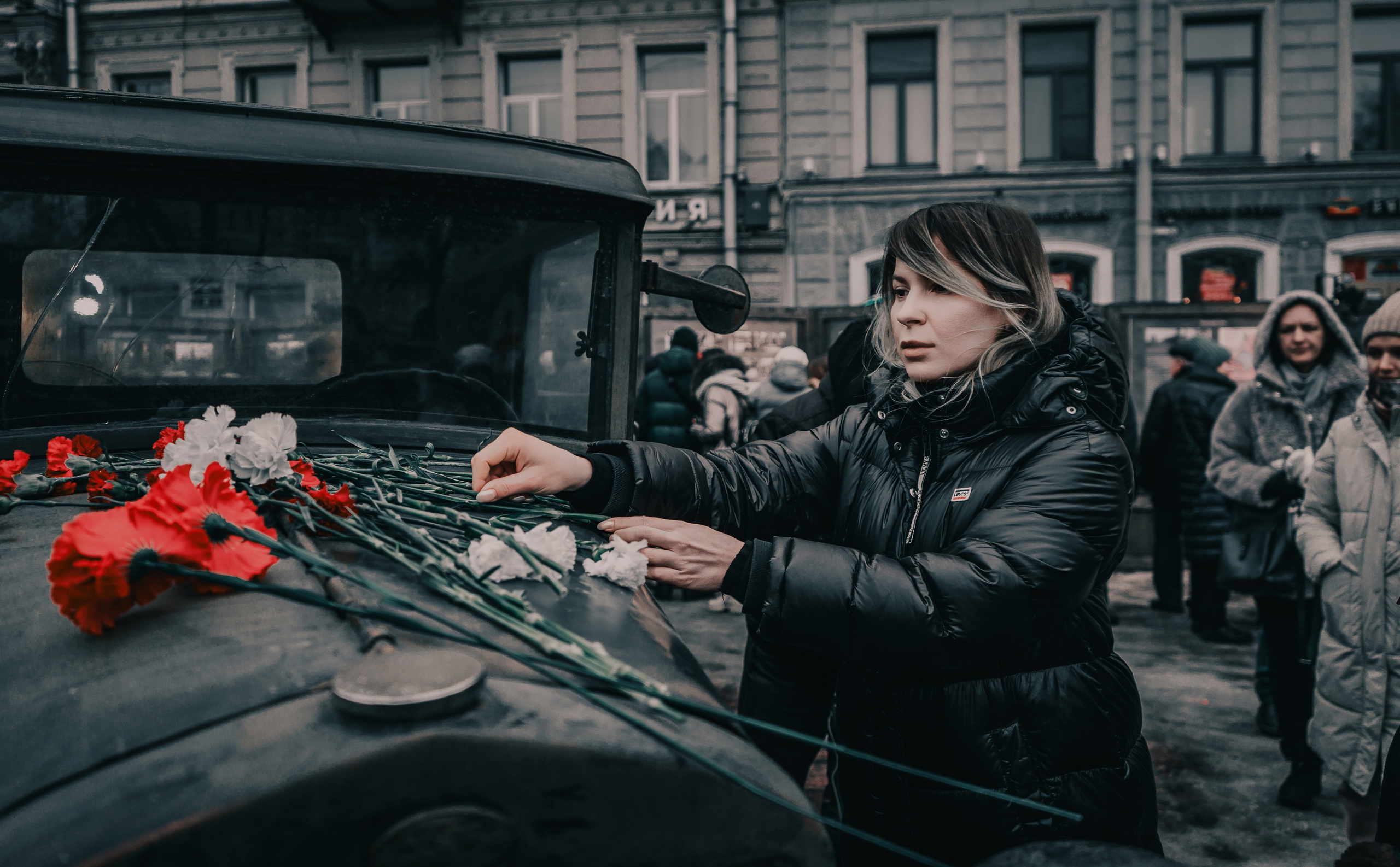 Женщина возлагает красные и белые гвоздики на капот старого грузовика на мемориальной акции. Уличная фотография Мальцева Евгения.