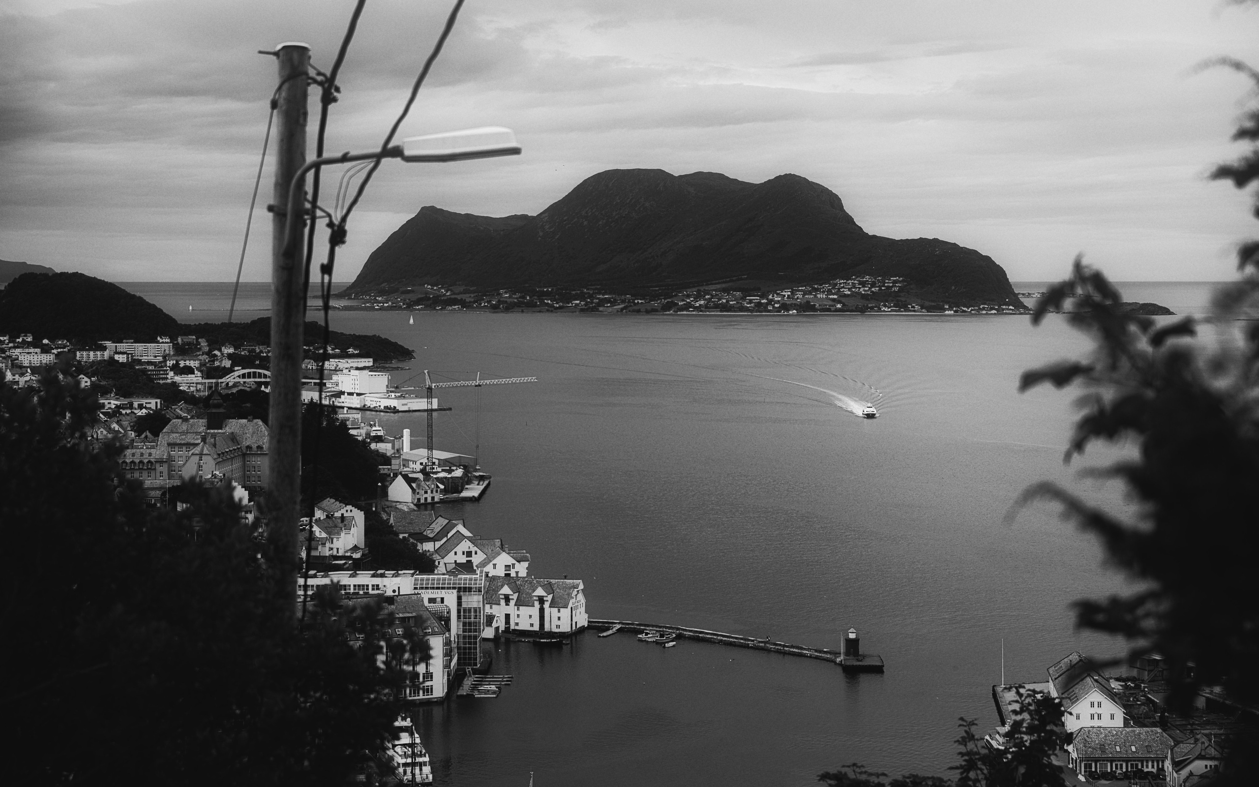 Черно-белая панорама: вид на прибрежный город и фьорд с высоты, лодка на воде, горы на фоне, уличный фонарь на переднем плане, Норвегия