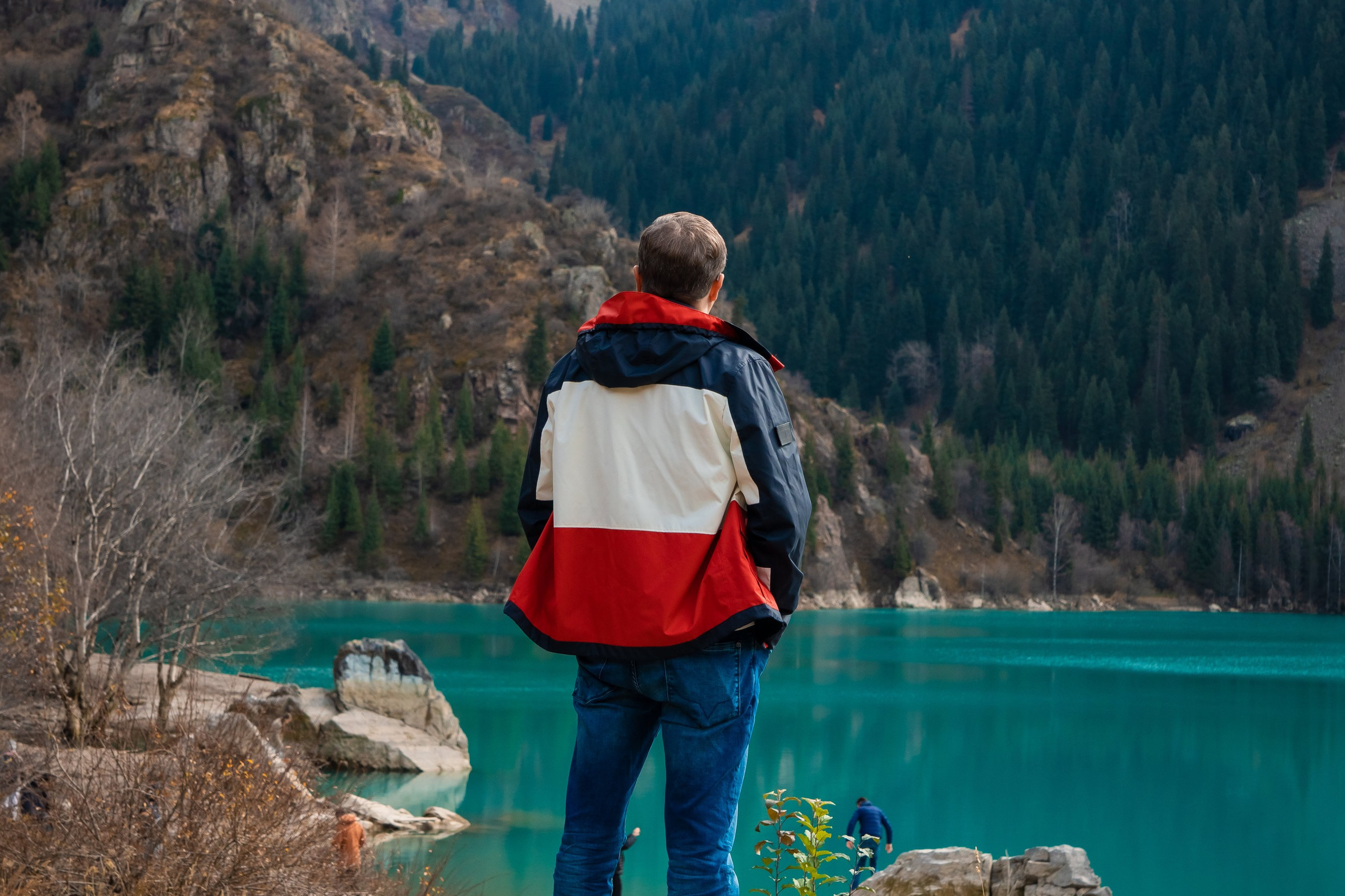 Горное озеро Иссык в Алматинской области, Казахстан, с бирюзовой водой на фоне осеннего леса с яркими золотистыми деревьями, окружённого высокими горами, покрытыми хвойным лесом. На горизонте видны заснеженные вершины под облачным небом