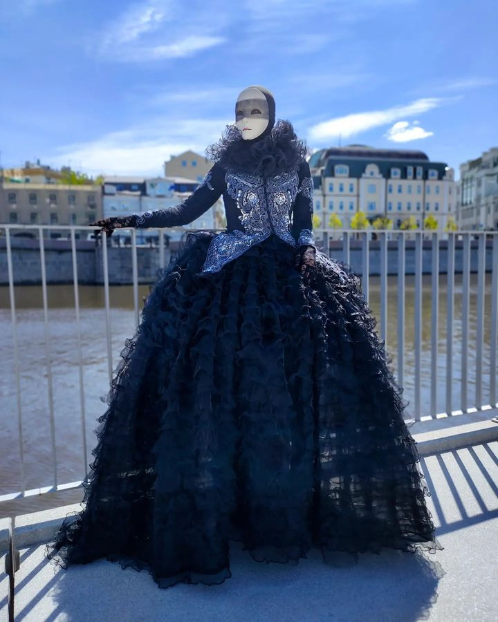 Артист в чёрном платье в белой маске стоит на набережной реки Москвы.