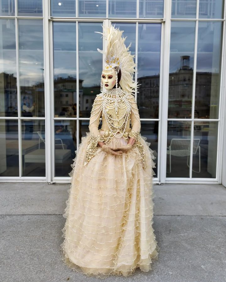 Мим в светлом белом костюме в маске в шапке с ирокезом на раусе у ресторана.