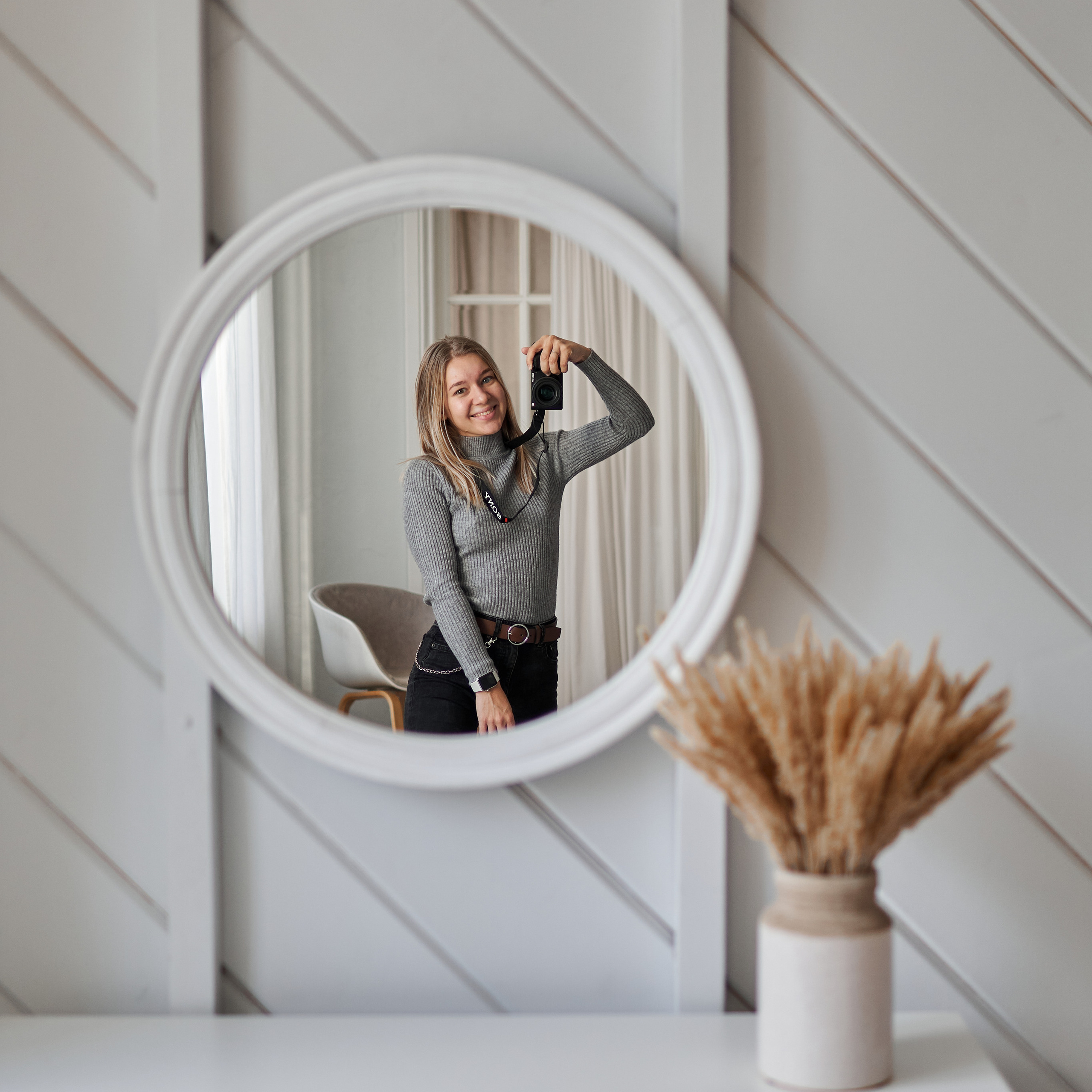Девушка с камерой в руках фотографируется в круглое зеркало, которое на белой деревянной стене