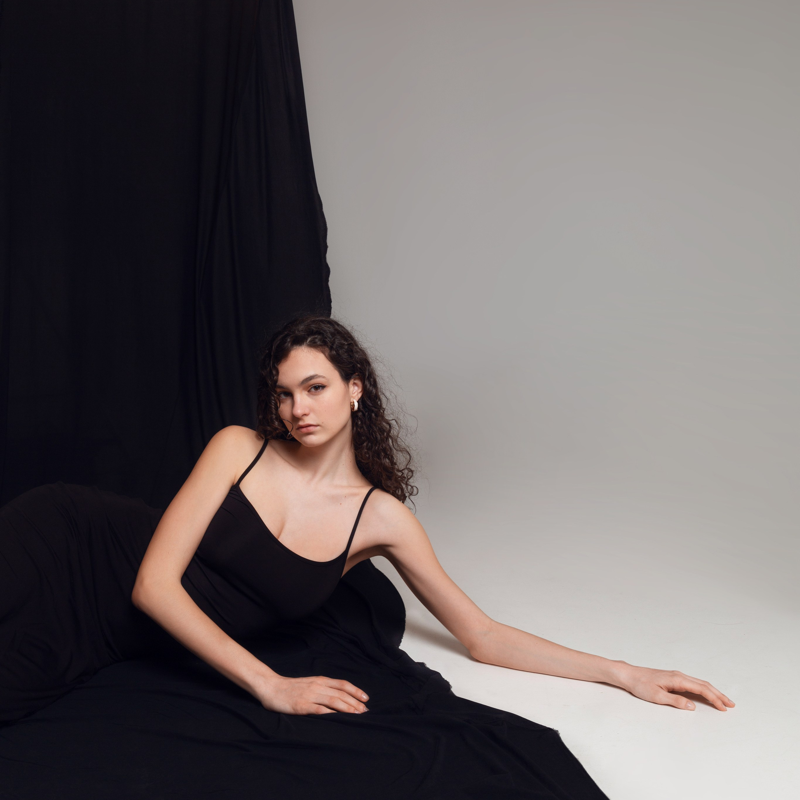 Девушка в длинном чёрном платье позирует на белом фоне. Пример профессиональной фотосессии в студии.