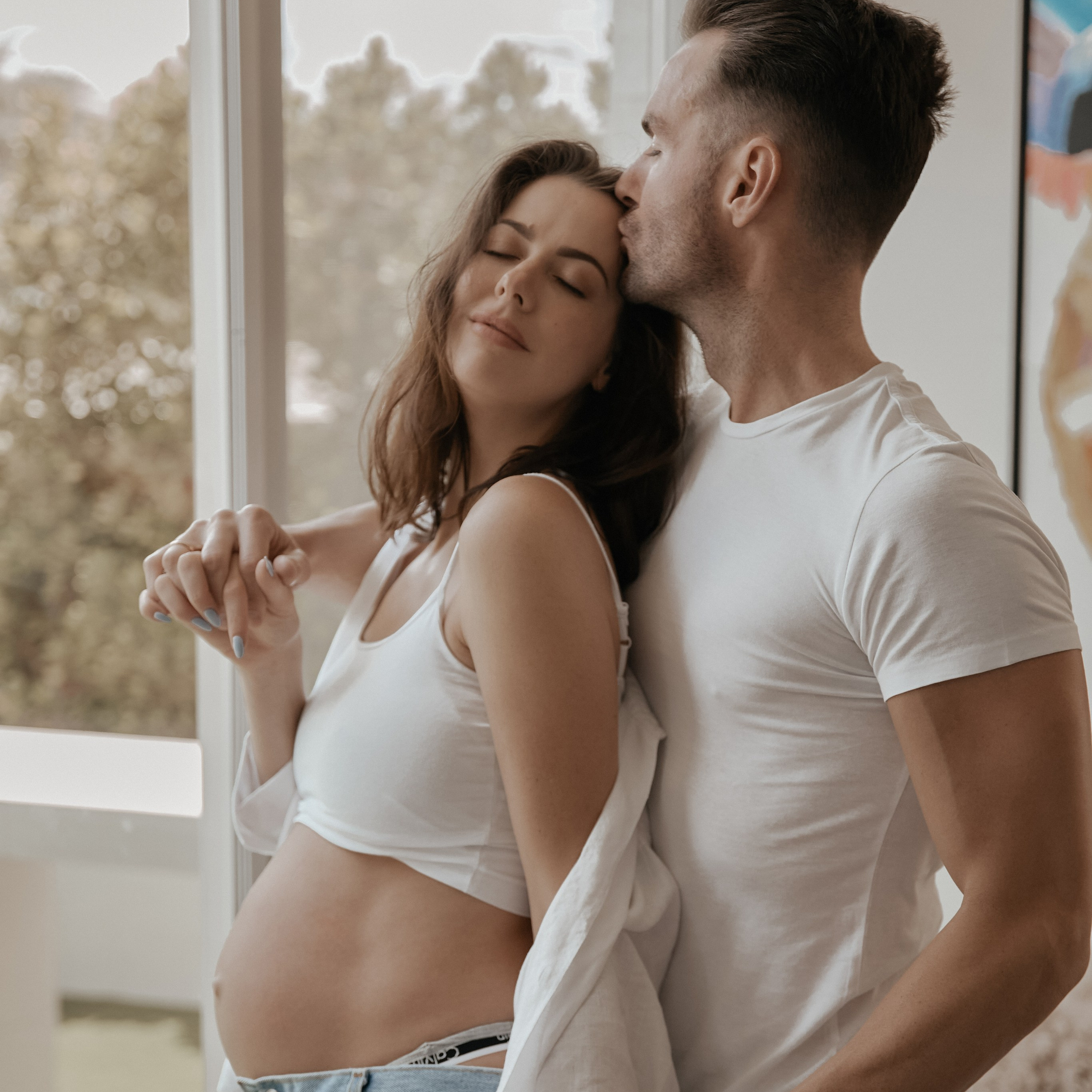 A pregnant woman leans against her partner, who kisses her forehead as they hold hands and stand by a window