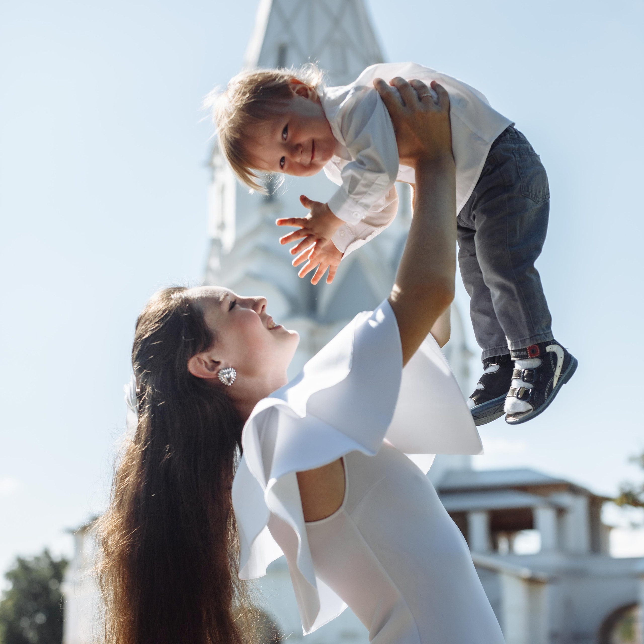 Мама с длинными шикарными распущенными волосами поднимает над собой ребёнка на вытянутые руки на фоне парка Коломенское, семейная фотосессия