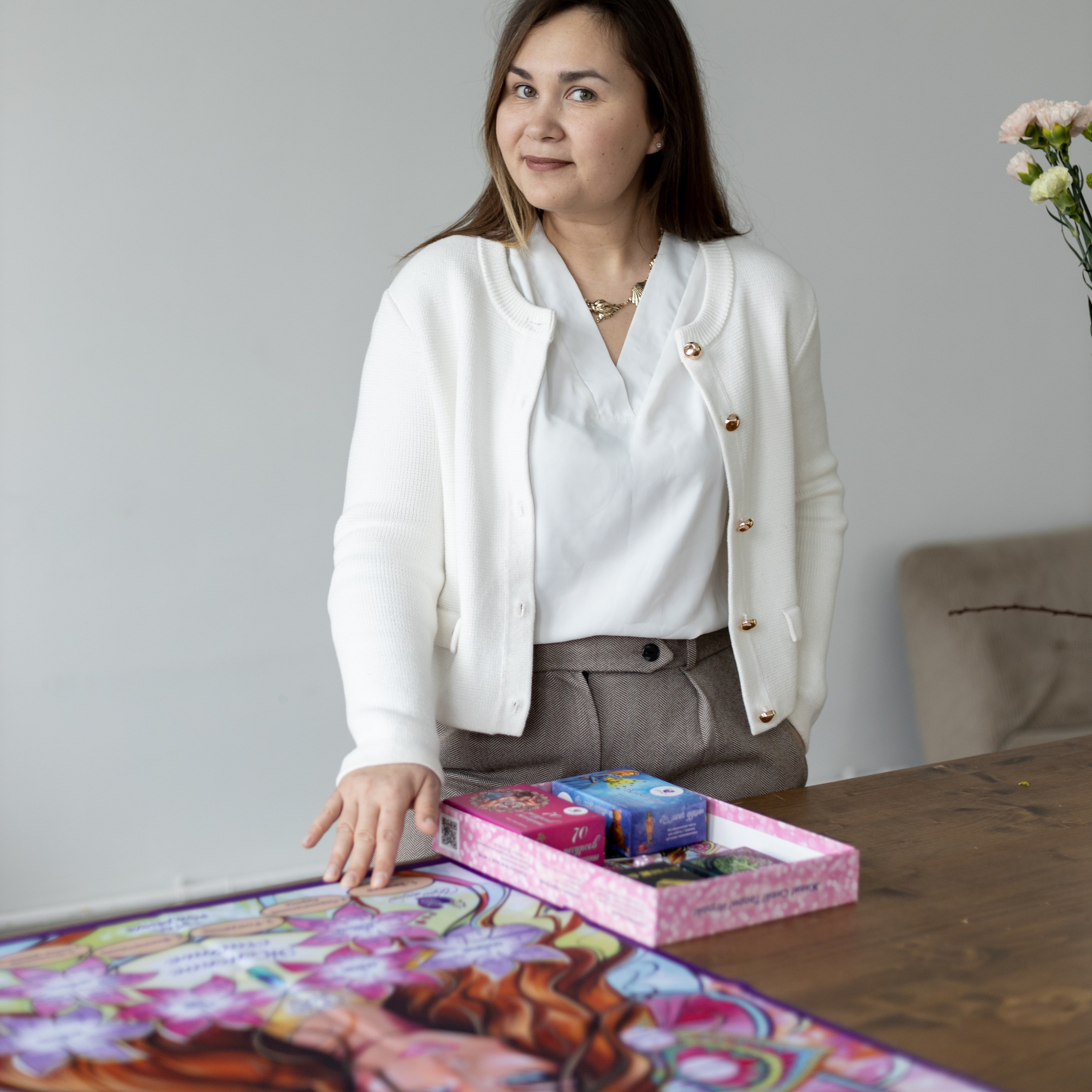 Юлия Андреева проводит трансформационную игру Женское счастье с женщиной за столом, на стульях разложены карты, участница внимательно слушает и выбирает ответ