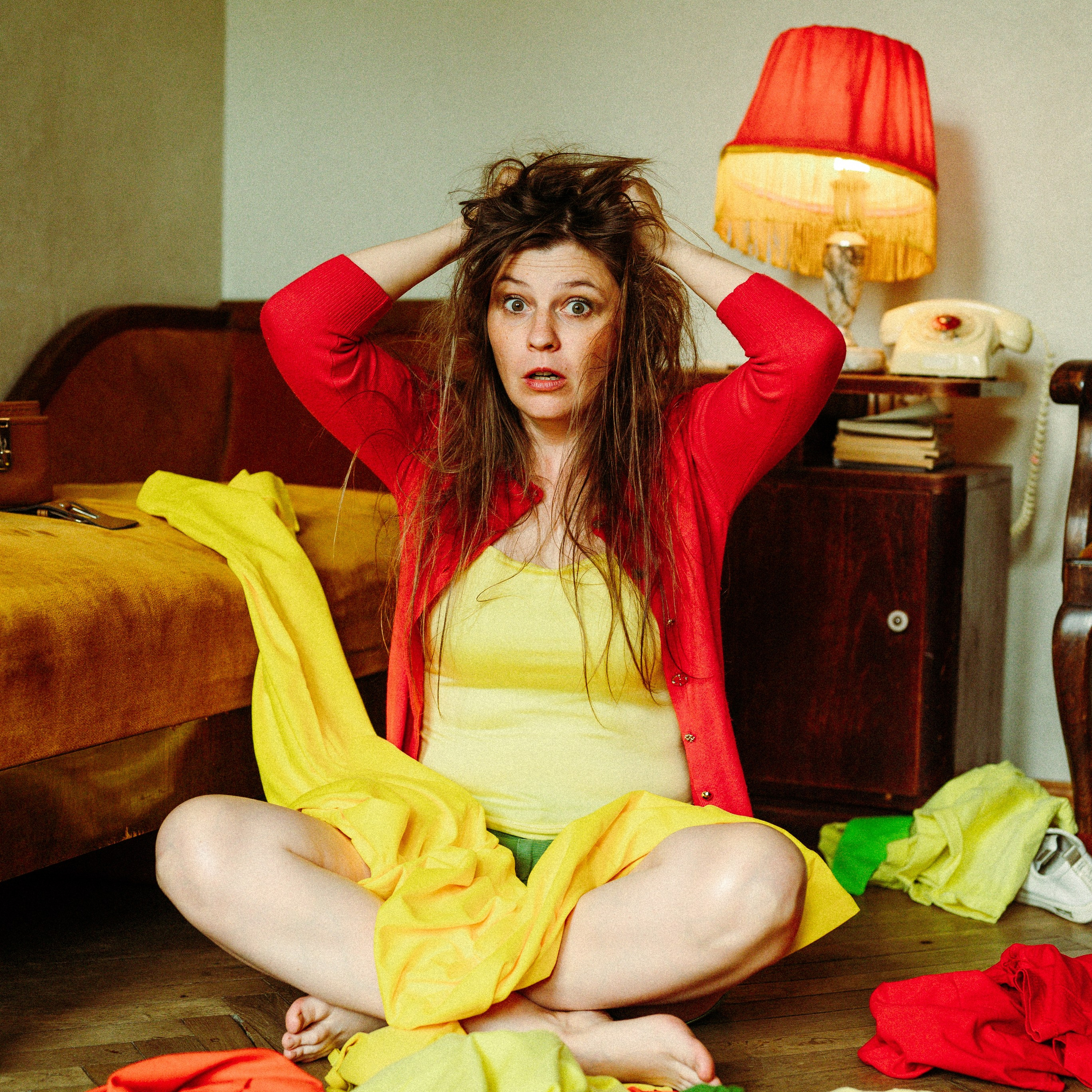 взъерошенная девушка сидит на полу среди разбросанных ярких вещей