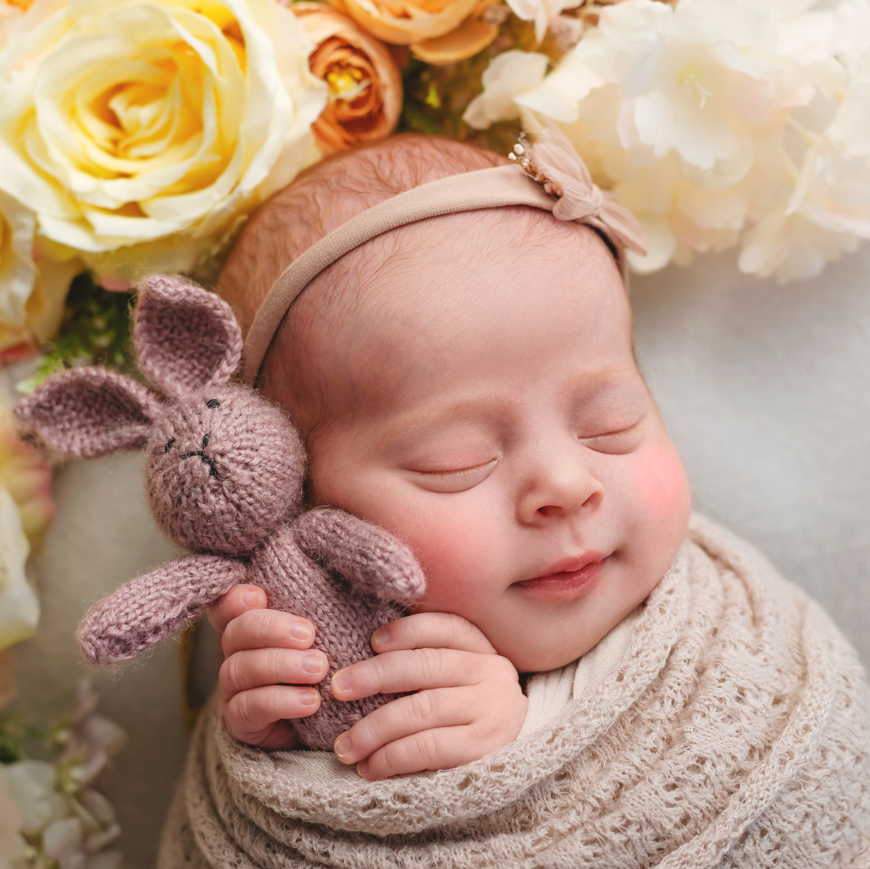 Новорожденная девочка в бежевой обмотке  лежит на белом плетенном диване на кружевной подушке. За спиной желтые розы. На голове повязка-бантик.